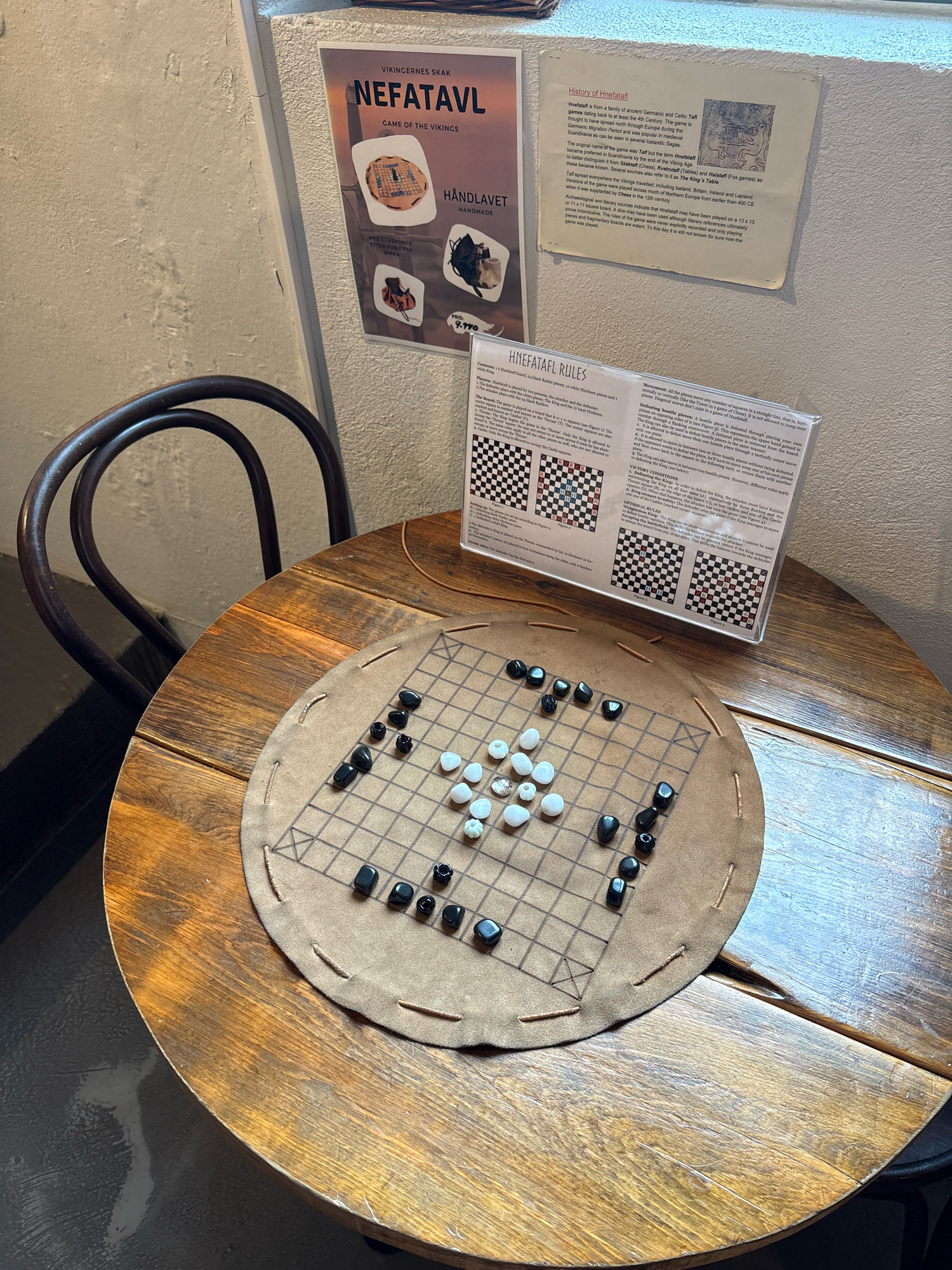 This image depicts a close-up view of a wooden table in what appears to be a cafe or a museum exhibit in Reykjavik, Iceland. On the table, a Hnefatafl game is set up on a round, tan mat with stitched edges. The game board is a grid with squares, and there are black and white playing pieces scattered on it, indicating an ongoing game. To the right of the game board is a clear plastic stand holding a folded sheet of paper with the rules of Hnefatafl, along with diagrams. Behind the table, a dark metal chair with a curved backrest is visible. On the wall in the background, there are two posters. The left poster has text in Swedish and images related to Viking games, featuring the game "Nefatavl". The right poster is a historical document or information card, with text and an illustration. The lighting suggests an indoor setting, possibly with ambient light from a window or overhead fixtures. The overall scene evokes a sense of history and strategy, likely part of a cultural display about Viking heritage.