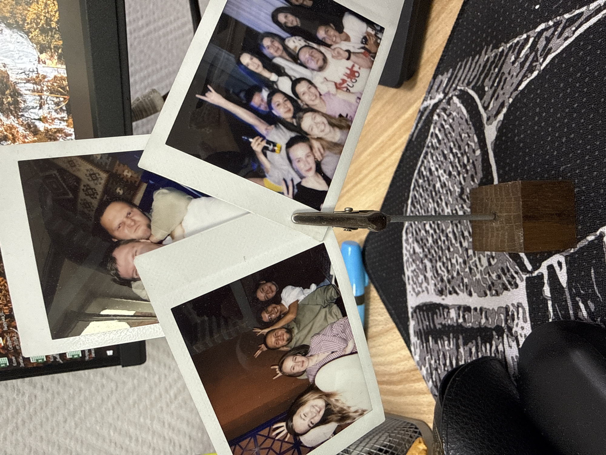 The scene shows a tabletop with several Polaroid photographs scattered across it. One photo displays a group of approximately ten women, with one holding a microphone and making a rock-on hand gesture. Another photo shows two men lying together, possibly on a patterned rug. A third photo features a group of around six women, all smiling and posing. A fourth photo shows a landscape, possibly a stream or waterfall. A metal clip attached to a wooden block holds the photographs together. A blue pen and a black patterned mousepad are also visible on the tabletop. The surface appears to be wood. The location, Krasnoyarsk, Russia, is not discernible from the image.