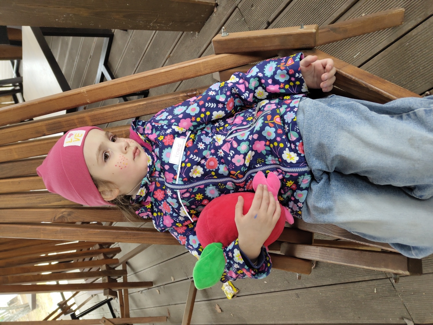 A child in a pink hat, floral jacket, and blue jeans lies on a wooden bench, clutching a red and green plush toy. The bench is outdoors, situated on a wooden deck, likely in an urban setting in Unknown, Unknown. Metal legs of tables and chairs are visible in the background.