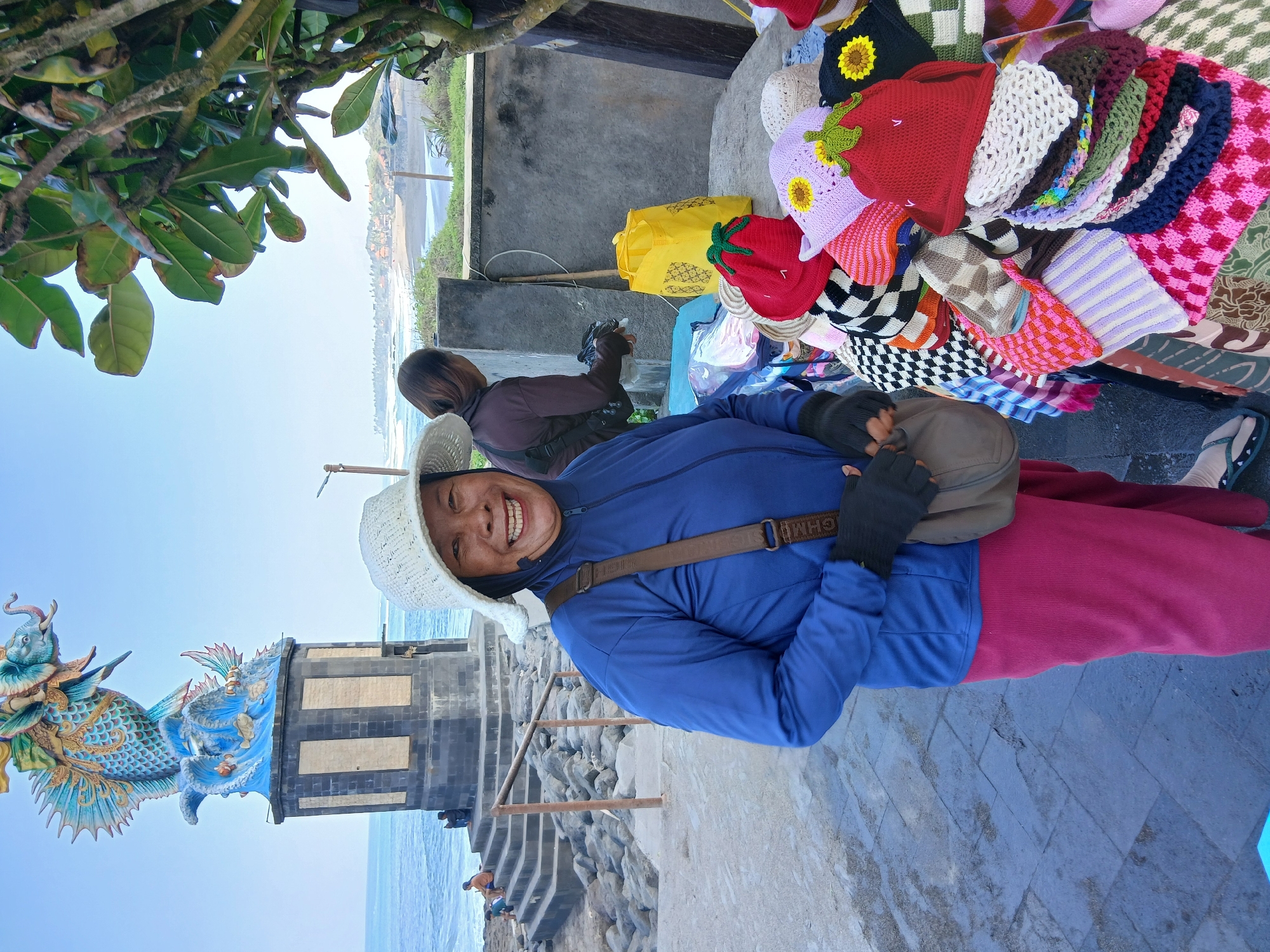 A medium shot captures a smiling woman in the foreground, wearing a white sun hat, a blue long-sleeved shirt, and dark gloves, with a brown crossbody bag. She stands on a paved walkway near a display of colorful knitted hats. Behind her, a larger statue of a mythical creature, possibly a dragon or a fish with a dragon's head, sits atop a stone pedestal by the ocean. The ocean is visible in the background, with waves crashing on a rocky shore and a sandy beach beyond. To the right, a woman in a dark shirt is partially visible, attending to the hat display. A bright yellow bag rests on a table laden with the knitted hats, which are varied in color and pattern, some featuring fruit or flower motifs. The environment appears to be a marketplace or tourist spot, likely near a beach or coastal area in Mengwi, Indonesia. The lighting suggests it is daytime, with a clear sky.