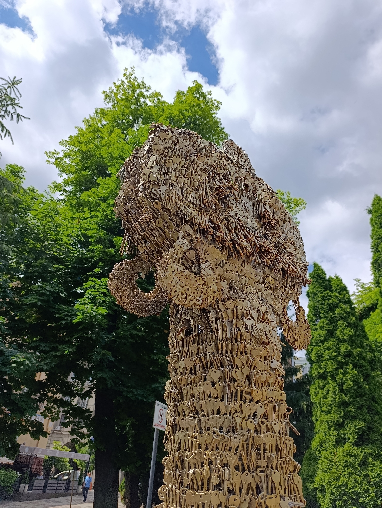 This image shows a close-up of a large sculpture made of many keys, possibly depicting an animal head with large ears. The sculpture is outdoors, with lush green trees and a cloudy blue sky in the background. In the distance, there is a street with buildings and some people. A sign with Cyrillic text and a prohibition symbol is visible on a pole near the sculpture. The sculpture is located in Kyiv, Ukraine, specifically at the Kyiv Pechersk Lavra, a historic Orthodox Christian monastery.