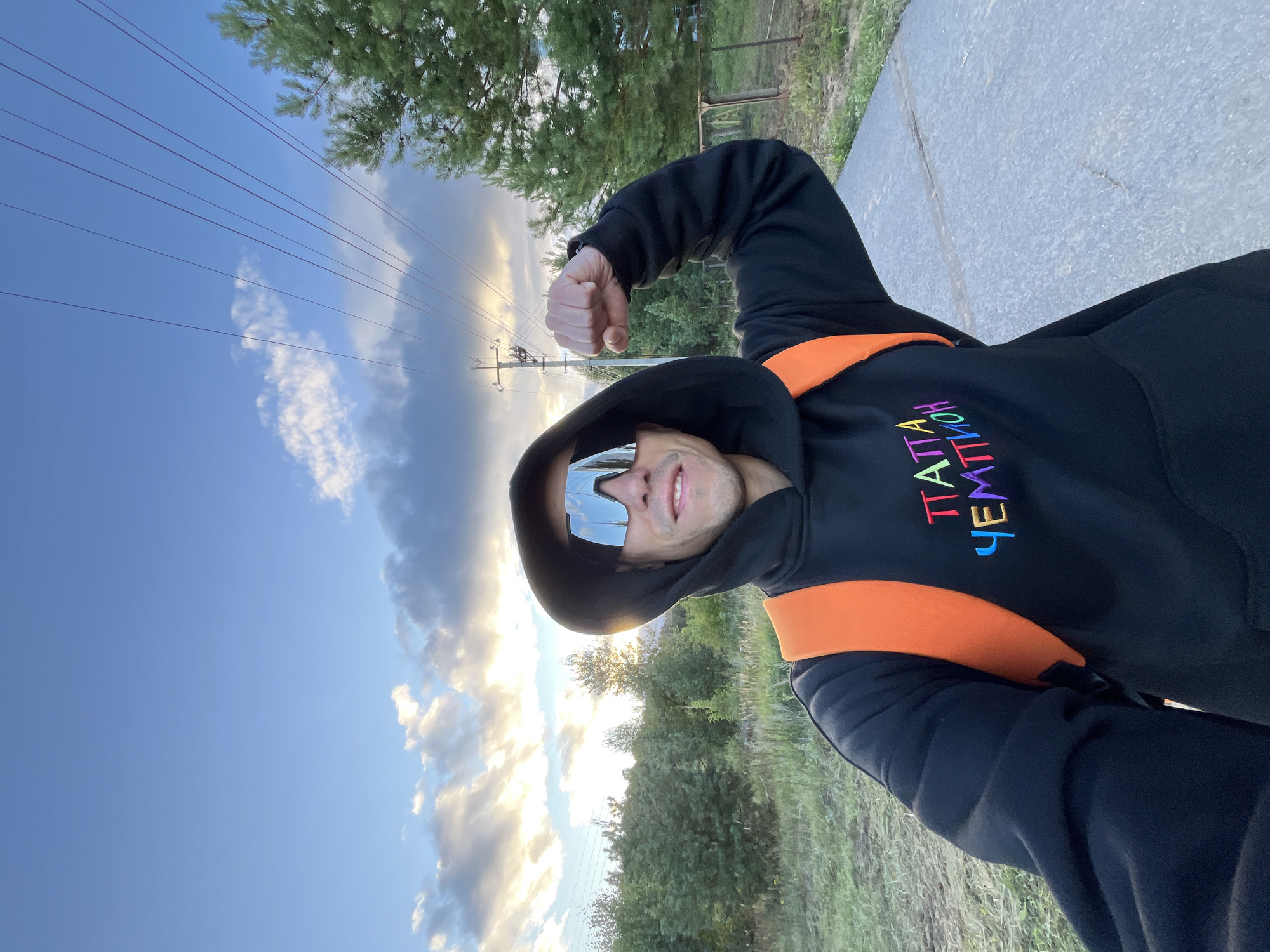 A man wearing a black hooded sweatshirt with an orange backpack and silver wrap-around sunglasses is posing for a selfie outdoors. The sweatshirt has colorful embroidered text on the chest. He is flexing his right arm and has a slight smile. The background shows a path, a fence, trees, and a cloudy sky with power lines overhead. The location is identified as Chaikovsky, Russia.