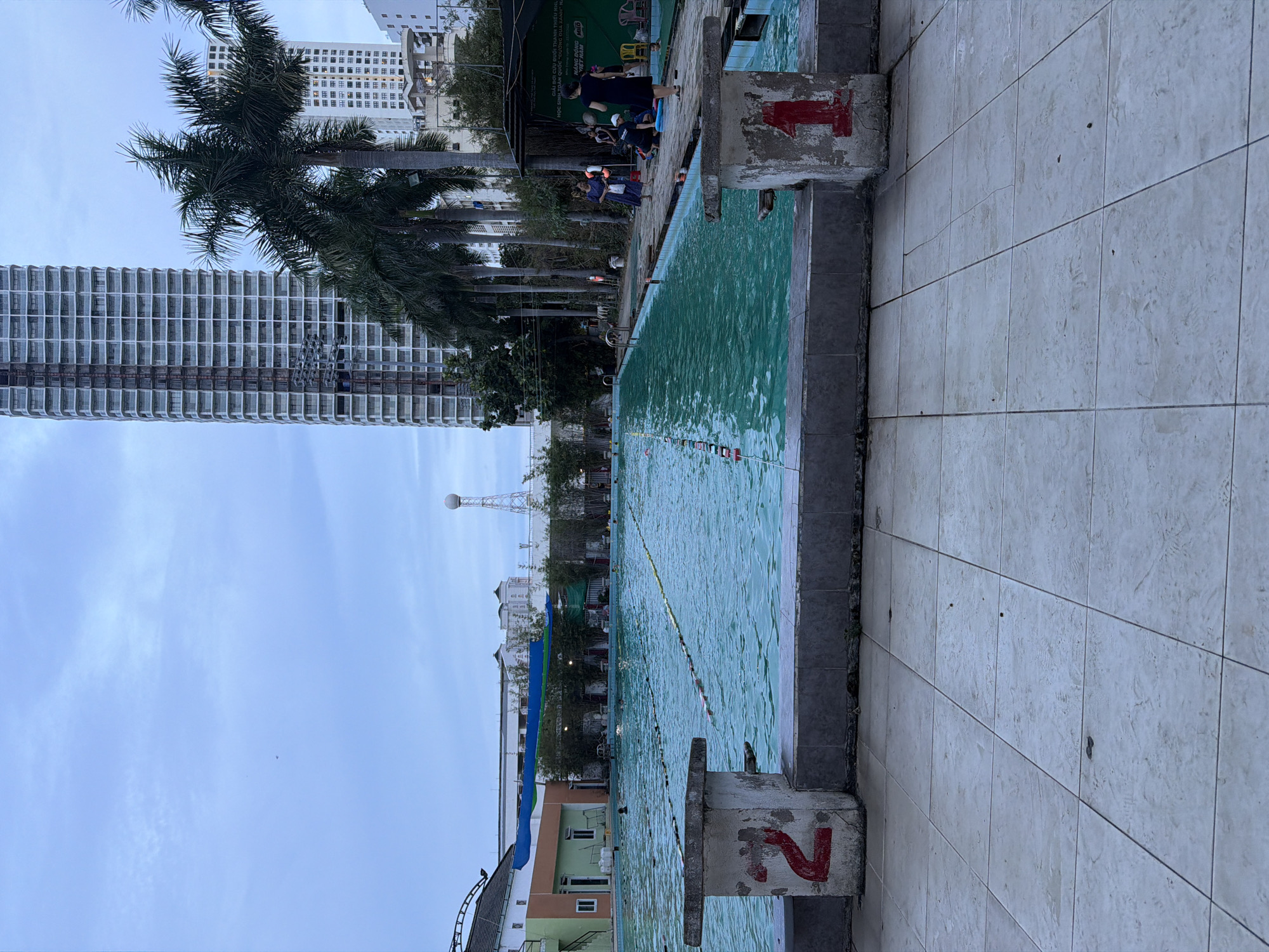 The image depicts a public swimming pool in Nha Trang Ward, Vietnam, with a cloudy sky overhead. The pool is a large, rectangular body of turquoise water, divided into lanes by floating markers. In the foreground, a tiled walkway surrounds the pool. On the left, a concrete structure with a red number "2" painted on it is visible. On the right, a similar concrete structure with a red number "1" is present.
Several people are scattered around the pool area. Some are in the water, swimming or wading, while others are on the poolside. On the right side of the image, a group of individuals are gathered near a covered area, with some sitting on chairs. Palm trees and other foliage line the edge of the pool. In the background, modern high-rise buildings dominate the skyline, contrasting with the more traditional architecture of the pool buildings. A water tower is also visible in the distance. The overall scene suggests a recreational area within an urban environment.
