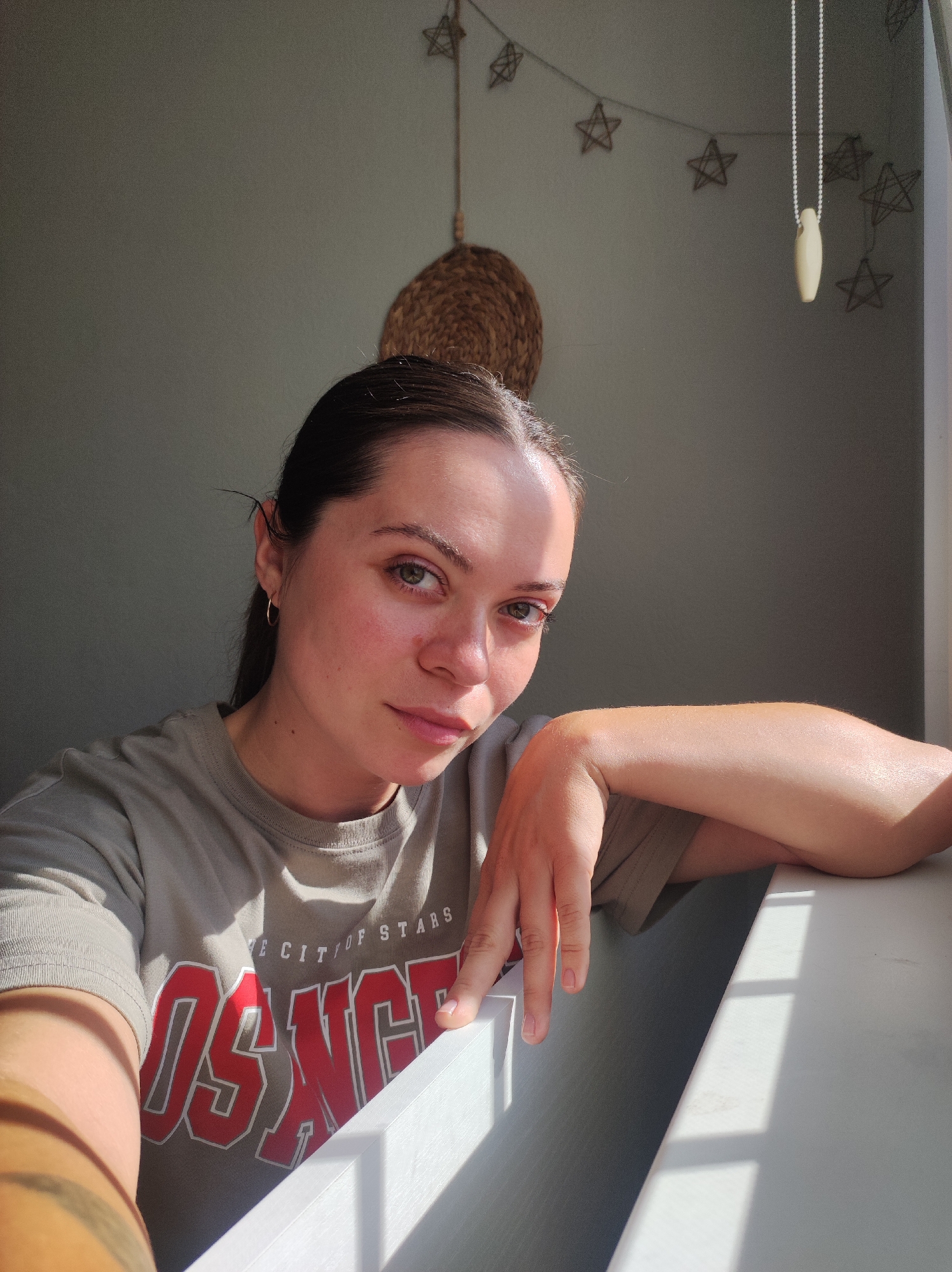 The image shows a young woman with brown hair pulled back, looking directly at the camera. She is wearing a light brown t-shirt with red lettering that appears to say "LOS ANGELES THE CITY OF STARS". She is leaning on a white surface, with her right arm resting on it. The scene is indoors, near a window, with sunlight casting shadows on the wall and the white surface. There are star-shaped decorations hanging on the wall above and to the right of the woman, and a woven circular object hanging on the wall to her left. The time of day appears to be daytime, likely late morning or early afternoon, given the strong sunlight. The woman's expression is neutral.