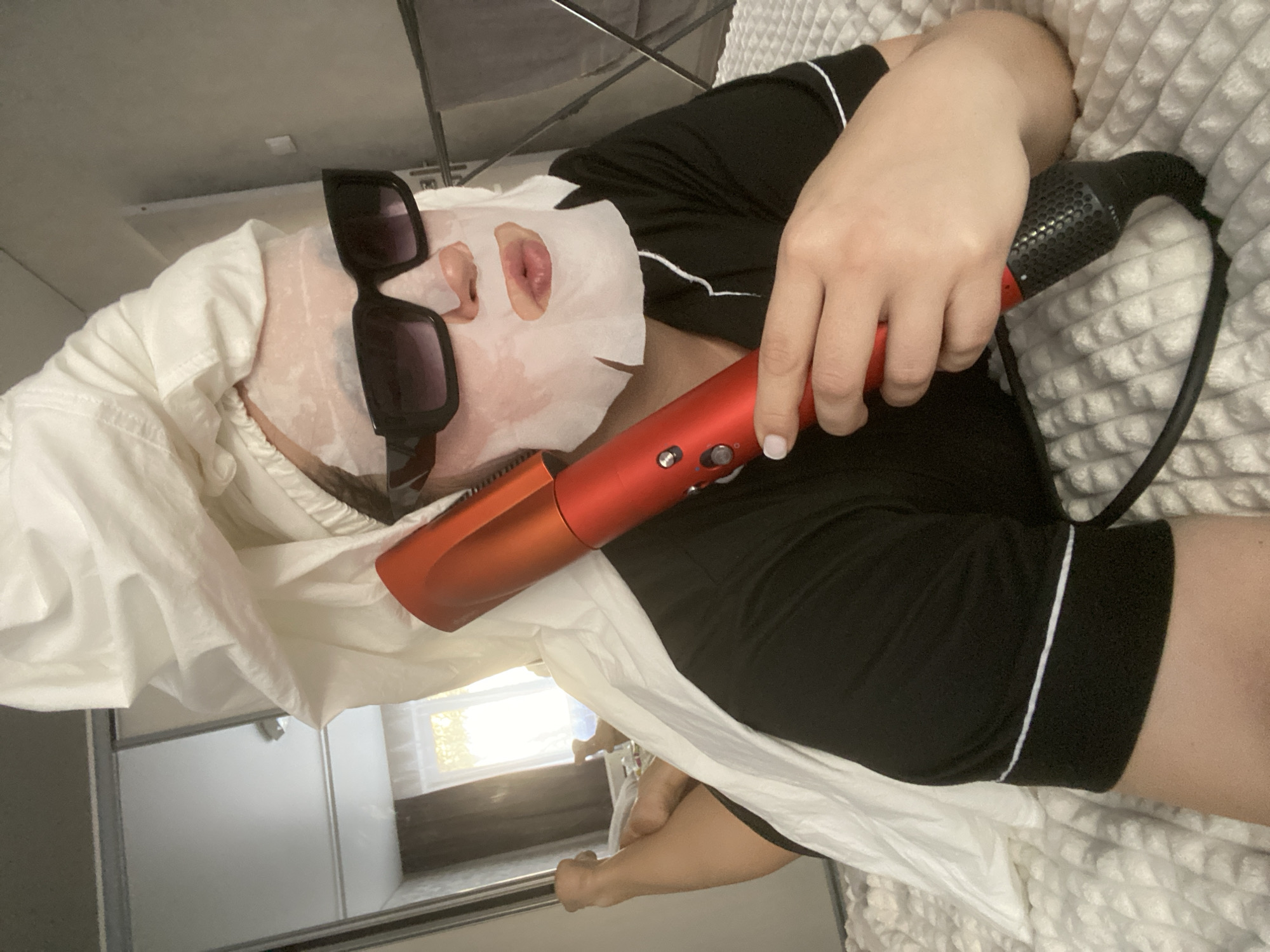 A person lies on a bed covered with a white textured blanket. They are wearing black pajamas with white piping, a white sheet draped over them, and black sunglasses. A white facial mask covers their face. They are holding a red hair dryer. The background includes a window, part of a shower or sliding door, and furniture. The location is not discernible from this image, and there's no visible evidence of Sochi, Russia.