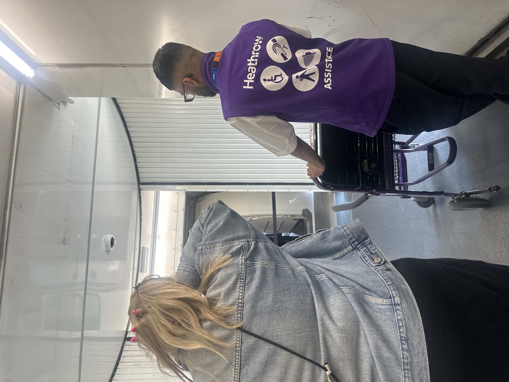 The image captures a scene inside an airport jet bridge, likely at Heathrow Airport in London, United Kingdom. A woman with blonde hair and a denim jacket is seen from behind, facing away from the camera. She has bright pink nails and a black strap from a bag across her shoulder. To her right, a man wearing a purple vest with "Heathrow ASSISTCE" and accessibility symbols printed on the back, is pushing a purple wheeled cart. The man is wearing glasses and appears to be an airport staff member providing assistance. The jet bridge is enclosed with white ribbed walls and illuminated by overhead fluorescent lights. The floor is a dark gray industrial surface. A small circular camera is visible on the ceiling of the jet bridge.