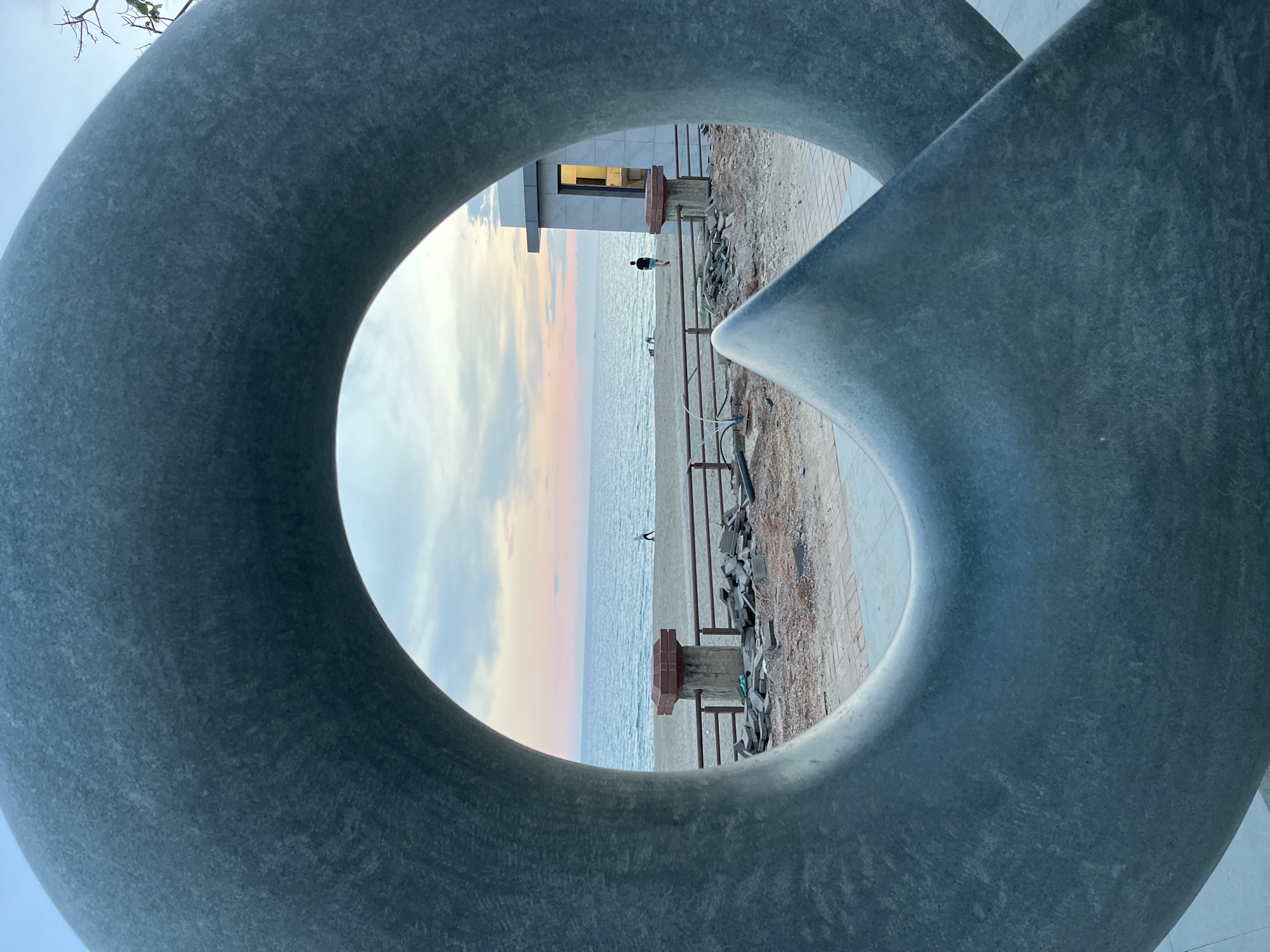The image captures a beach scene in Alanya, Turkey, viewed through a large, gray, abstract sculpture with a circular opening. The sculpture is the primary foreground element. Through the opening, a beach is visible, with a person walking near the water. To the left of the beach, there is a building. Above the sea, the sky shows a gradient of colors, indicating either sunrise or sunset. There are construction materials behind a metal railing. The walkway appears to be stone.