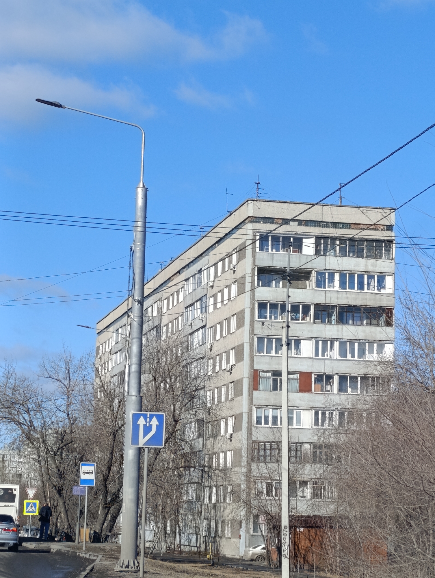 The image shows a gray, multi-story residential building in Krasnoyarsk, Russia, under a clear blue sky with scattered clouds. The building has rows of rectangular windows, some with visible air conditioning units. A gray lamppost stands in the foreground, with various road signs attached to it. Bare trees and branches frame the lower portion of the view. A road runs along the bottom edge, with a pedestrian crossing and a parked car visible. A person is standing near the crosswalk. Overhead electrical wires are stretched across the sky.