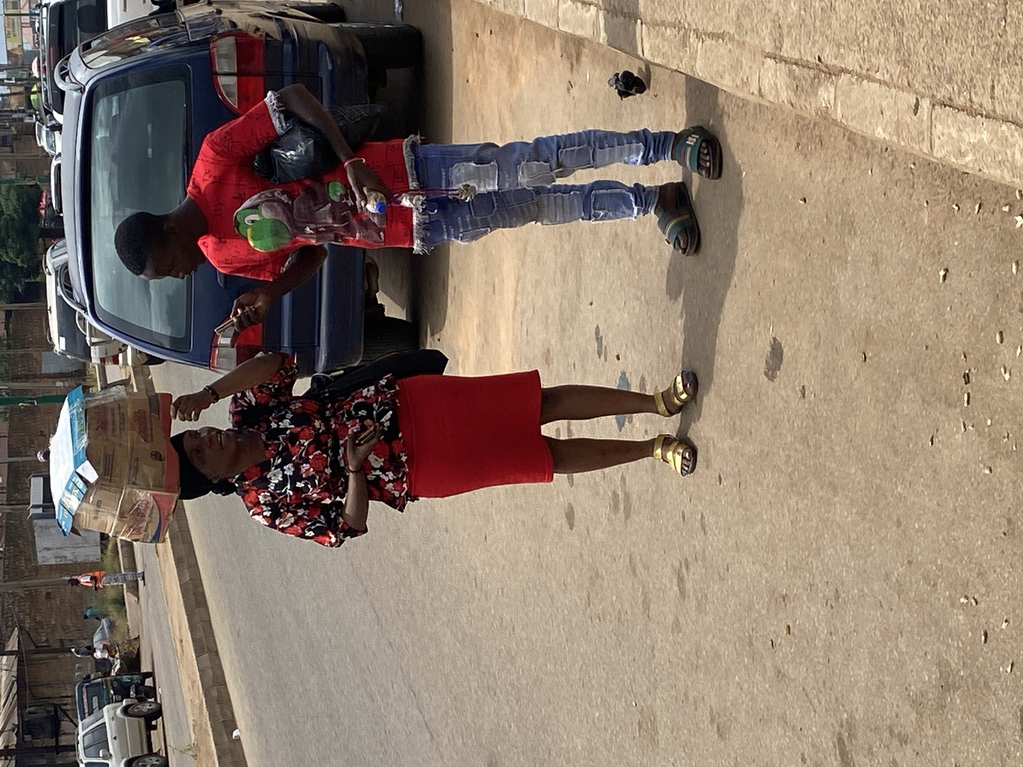 In Bukuru, Nigeria, a young man in a red t-shirt with a graphic print and ripped blue jeans stands next to a dark blue car, looking at his phone. He wears sandals. To his right, a woman carries a stack of boxes on her head, dressed in a floral-patterned shirt and a red skirt, also wearing sandals. The scene appears to be outdoors on a paved surface, possibly a roadside, under natural light suggesting daytime. The overall atmosphere is that of an ordinary day with people engaged in everyday activities.