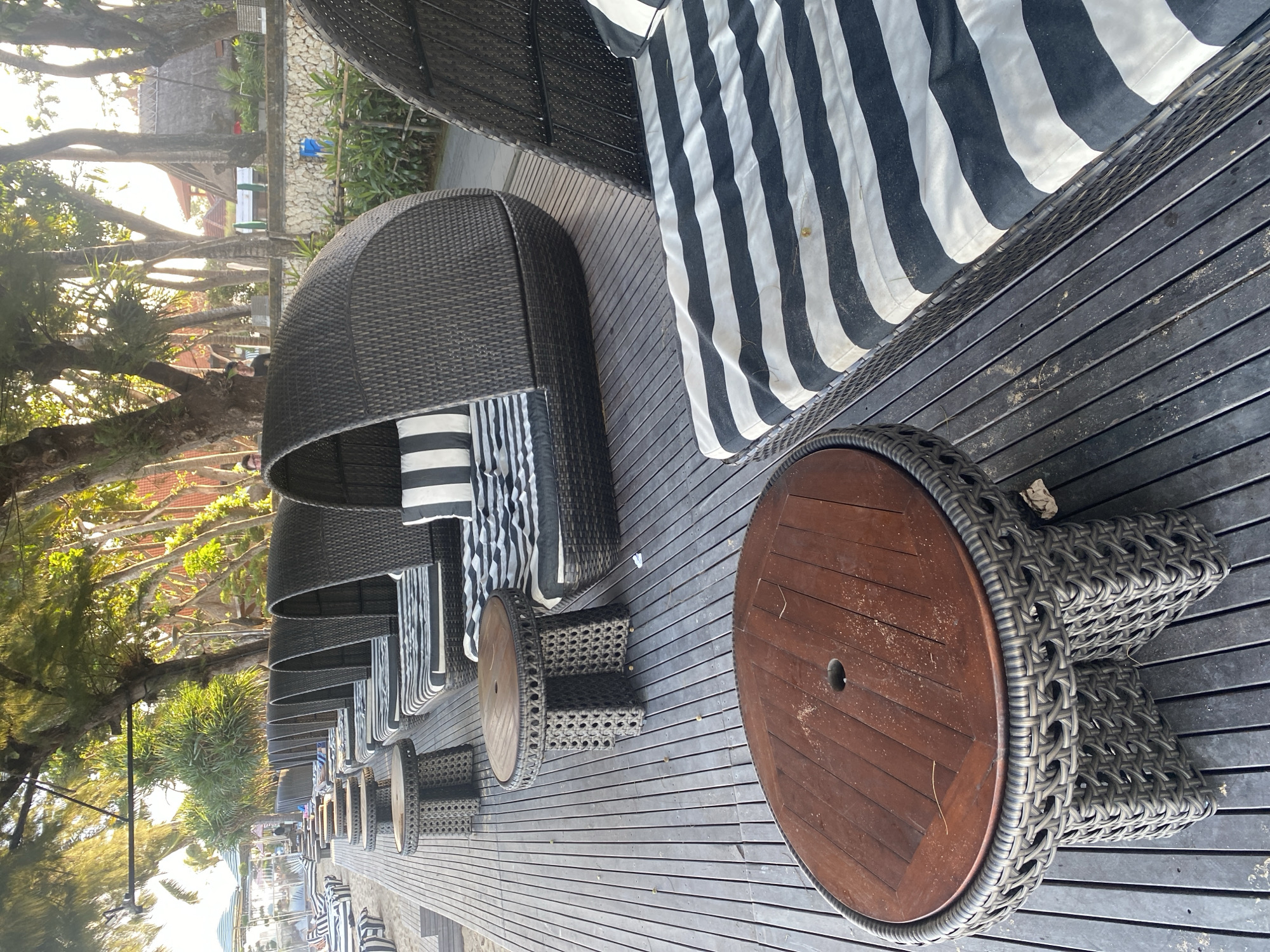 This image depicts a row of outdoor lounge chairs and small tables situated on a wooden deck. The lounge chairs are black, woven wicker with striped black and white cushions, and some have hoods providing shade. The tables are also made of woven wicker with round, wooden tops. In the foreground, a larger wooden table is visible. The deck is laid out on a beach or near a body of water, with sand visible on the left side of the image. Lush green trees with dappled sunlight filtering through their leaves line the background, suggesting a tropical environment. In the distance, there are glimpses of buildings with thatched roofs and some smaller lounge chairs and umbrellas, indicating a resort or hotel setting. The location is identified as Denpasar, Indonesia. The scene appears to be unoccupied at the moment captured in the photograph.