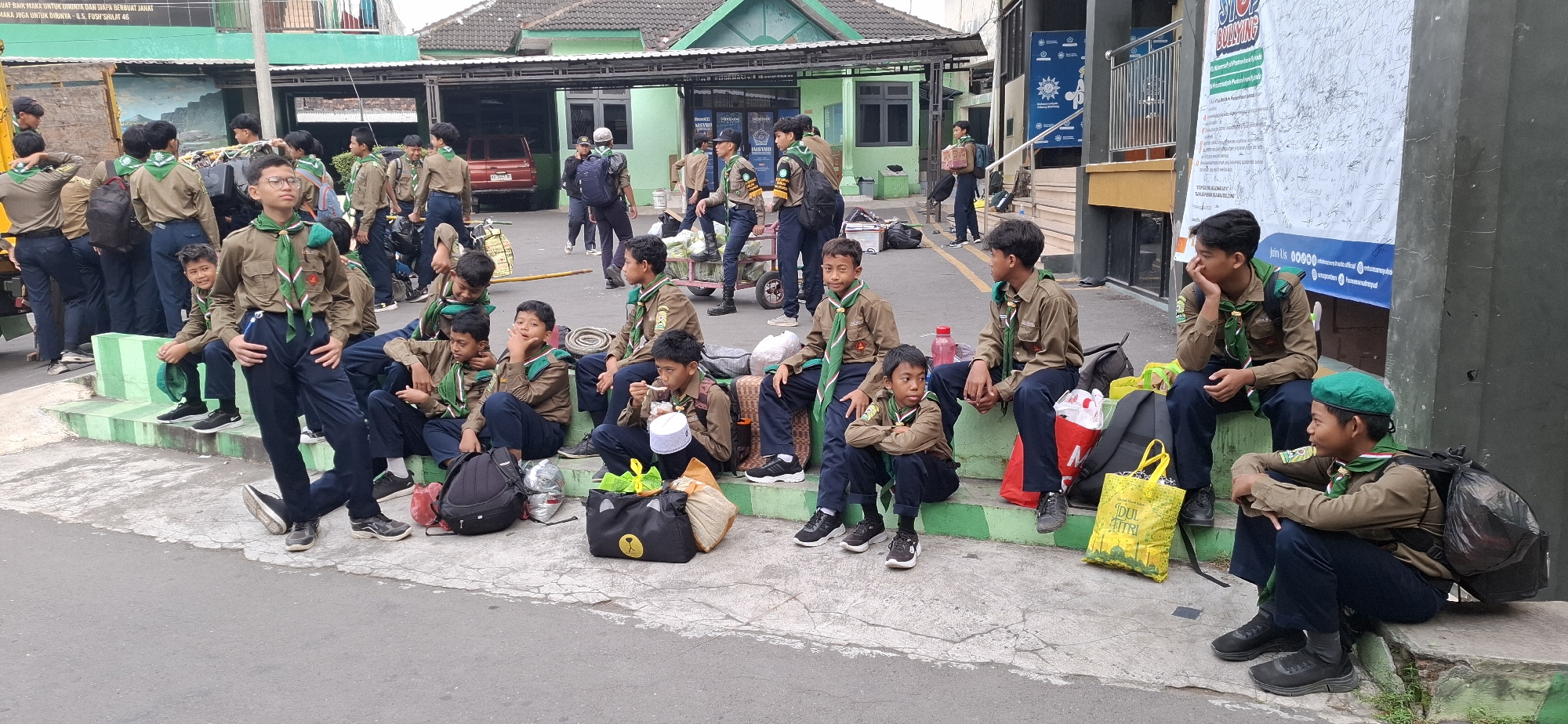 A group of young boys, dressed in scout uniforms consisting of khaki shirts, dark pants, and green neckerchiefs, are gathered outdoors. They are sitting and standing on concrete steps and the ground in front of a building with green trim. Some of the boys are looking in different directions, while others are interacting with each other or engaged in individual activities. Bags, including backpacks and tote bags, are scattered around them. In the background, a red car is visible, along with a building structure and trees. The scene appears to be a public area in a city, possibly a school or community center.