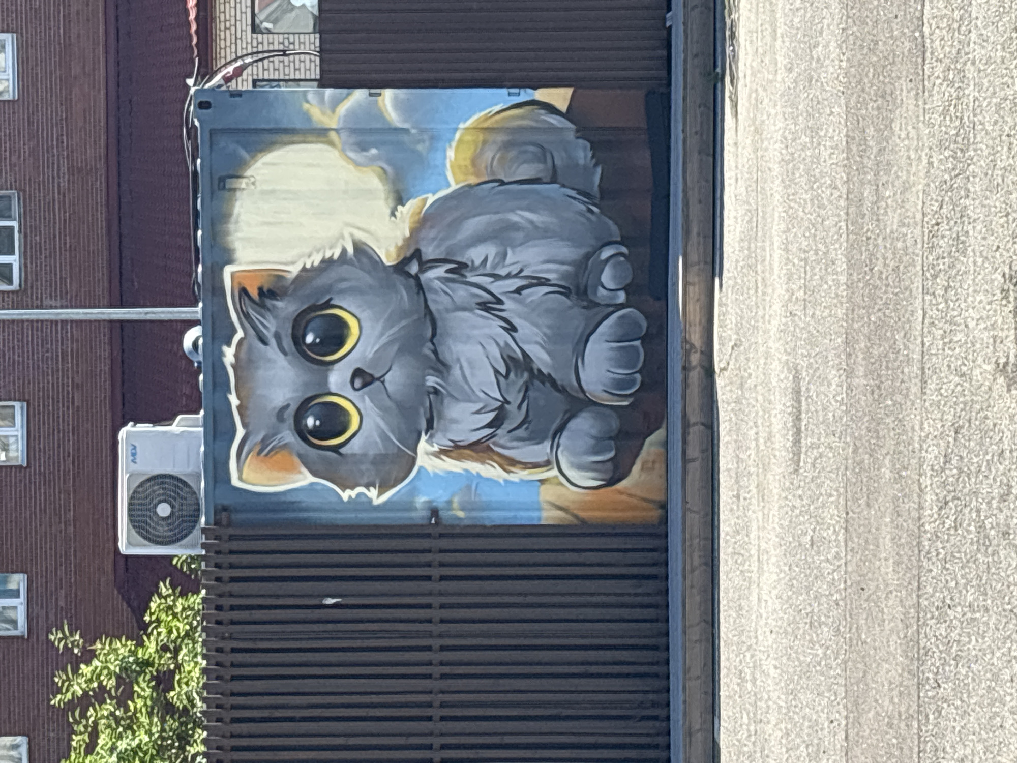 A mural of a large, fluffy, grey cat with big yellow eyes is painted on the side of a shipping container. The cat is depicted sitting down, with its paws tucked underneath it. Behind the cat, a yellow moon and clouds are visible in a blue sky. To the left of the container is a dark brown wooden fence. Above the fence, an outdoor air conditioning unit is mounted on the side of a brick building. The scene is outdoors, with a paved area in the foreground.