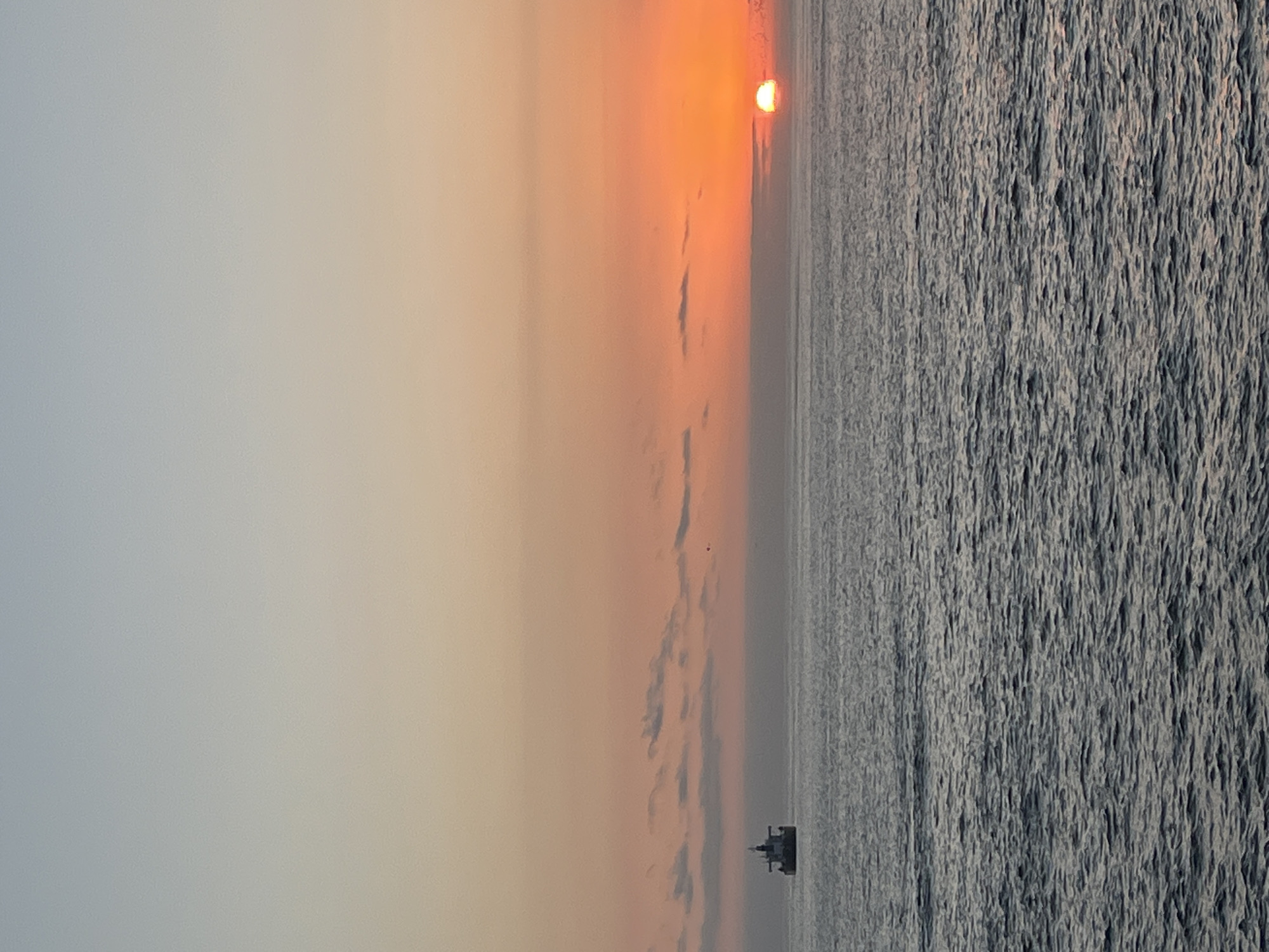 The image depicts a sunset over the Pacific Ocean, likely from the coastal city of Arica, Chile. The sun is partially obscured by the horizon, casting an orange glow in the sky. The sea appears choppy with defined wave patterns. A ship is visible on the horizon. The sky transitions from grey to orange towards the sunset.