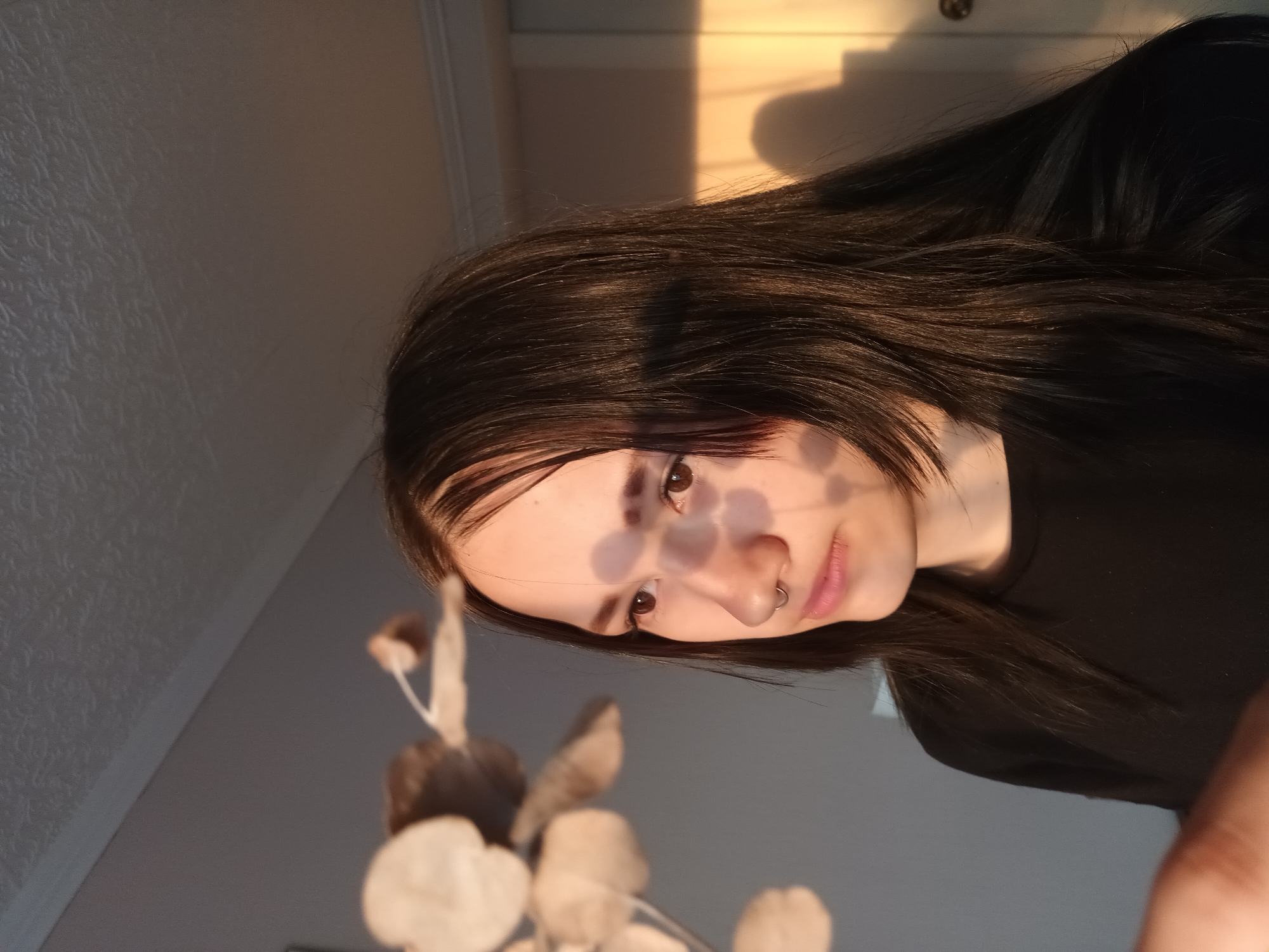 A young woman with dark hair and a nose ring is partially obscured by patterned shadows on her face. She is wearing a black shirt. Behind her is a gray wall with a hanging decoration of artificial leaves. The top right of the frame shows a light-colored wall and the shadow of an object, possibly a window frame. The location is unspecified, and the connection to Ольховка, Russia, is unsupported by the visual evidence.