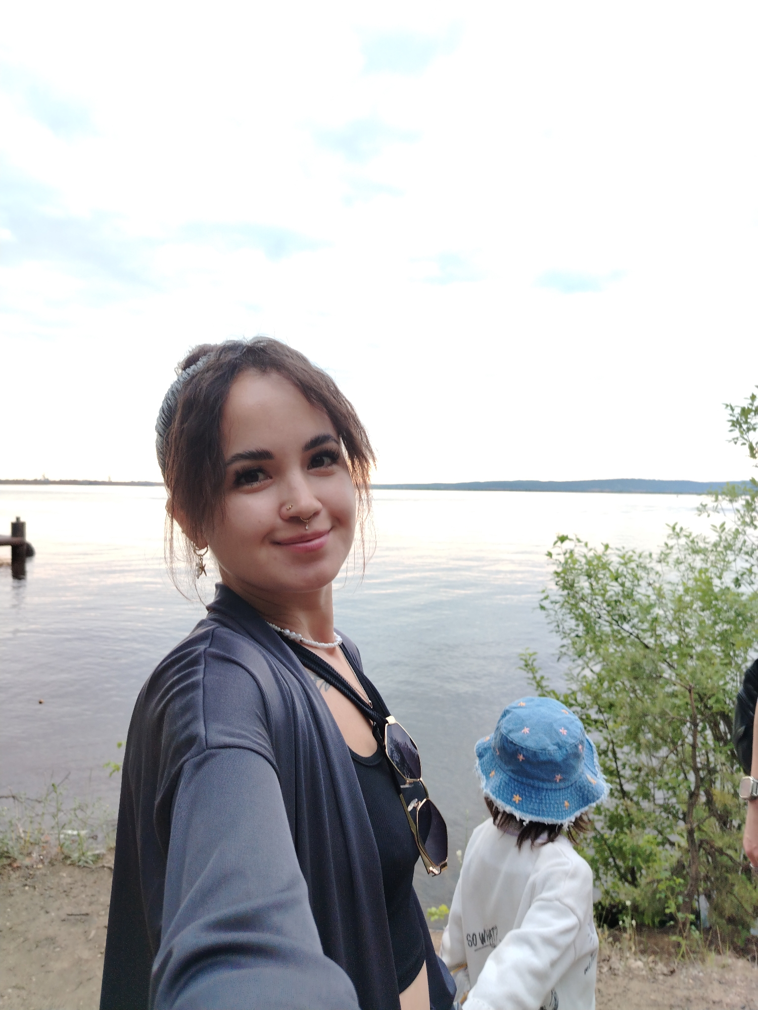 In Chaikovsky, Russia, a woman with dark hair pulled back is taking a selfie by a body of water, possibly the Kama River or a reservoir. She is wearing a dark gray open cardigan over a black top, a pearl necklace, and has hoop earrings. Behind her, to the right, a child wearing a blue bucket hat with orange floral patterns and a white jacket with the text "GO WHAT?" is facing away from the camera. The background features a calm, wide expanse of water under a cloudy sky, with a distant tree line and a wooden pier visible to the left. The ground in the foreground is a sandy, dirt path.