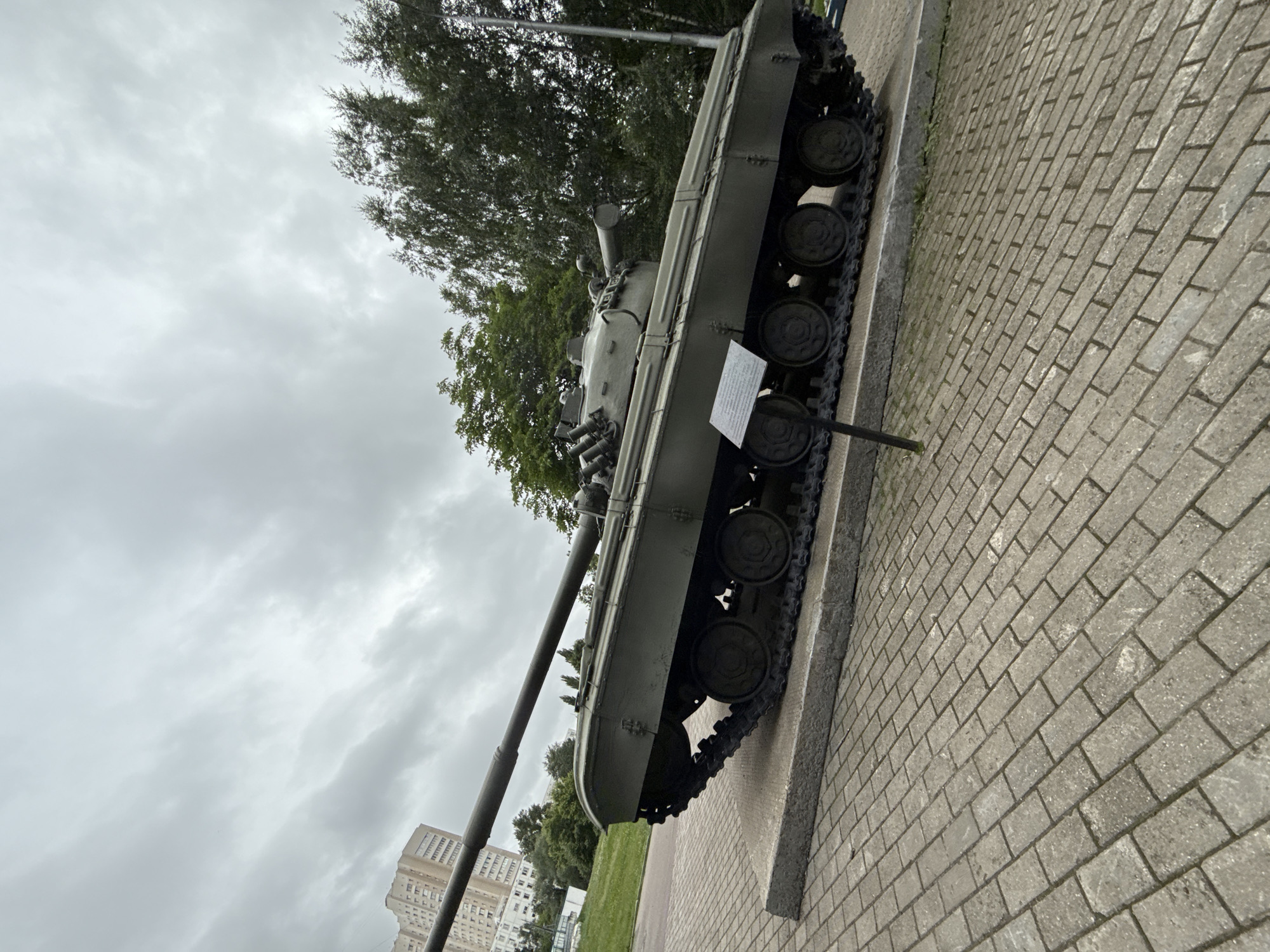 The image shows a parked military tank on display. The tank is olive green and appears to be an older model. It is situated on a concrete platform that is surrounded by a paved area. Behind the tank, there are trees and a tall building in the background. The sky is overcast and cloudy. The location is identified as the Panteleimonovskaya Church in Moscow, Russia.