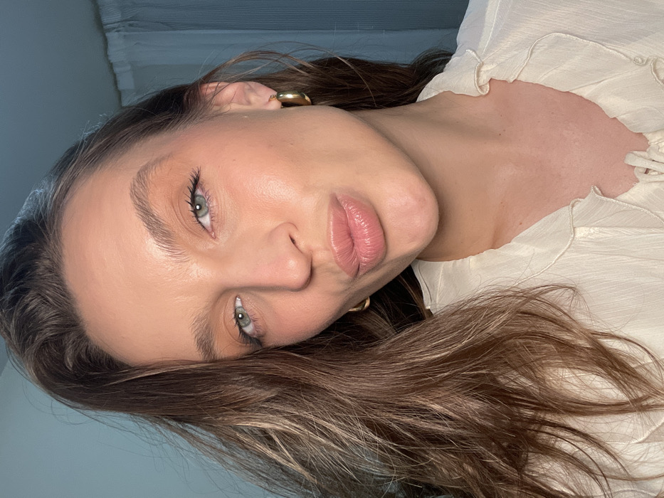 A woman with light skin and green eyes gazes at the camera. Her brown hair with blonde highlights is casually styled. She wears gold hoop earrings and a white, ruffled blouse. Her lips are full and slightly pursed. The lighting accentuates her makeup, including defined eyebrows and a dewy complexion. The background is muted, focusing attention on her face and upper body.