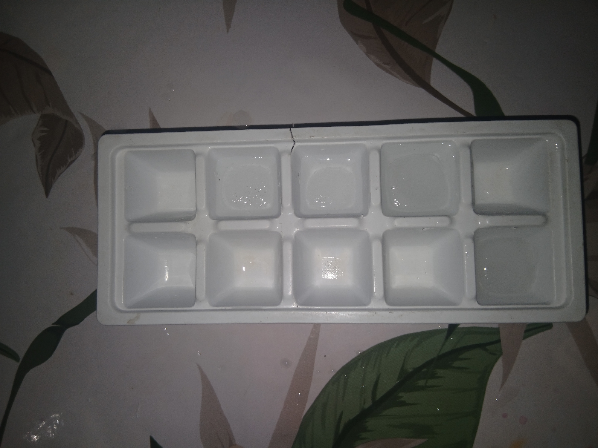 An overhead shot shows a white plastic ice cube tray on a patterned surface. The tray contains 12 square compartments, some of which are partially filled with clear liquid, possibly water or ice. The patterned surface appears to be a tablecloth or placemat with stylized leaf designs in shades of green and brown. The lighting is dim, casting shadows and highlighting the reflections in the liquid. There is a small crack on the right side of the ice cube tray. The location is identified as Parkent, Uzbekistan.