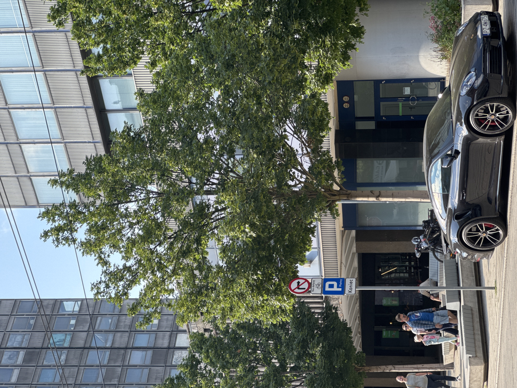 In the city of Poznan, Poland, on a sunny day, a dark-colored sports car is parked on the street in front of a modern office building with large windows. Trees line the sidewalk, providing shade. A traffic sign indicates no right turn and that parking is paid. Several people are walking and standing on the sidewalk, some with children. A bicycle is parked near the building entrance. The scene appears to be in a commercial or urban area.
