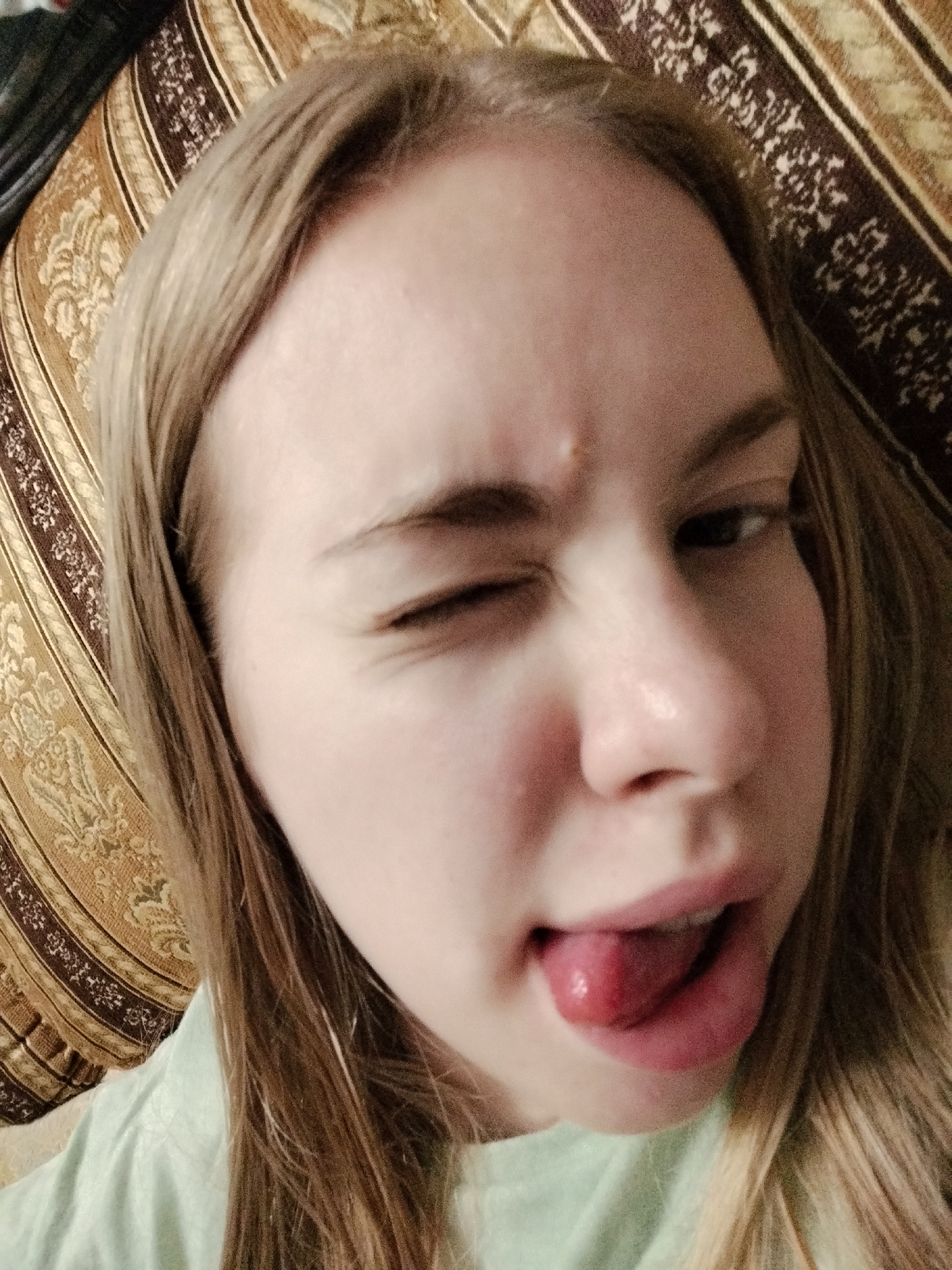 The image is a close-up, high-angle selfie of a young person with blonde hair, wearing a light green shirt. The person is winking with their right eye and sticking their tongue out. They are positioned in front of a patterned cushion with beige and brown stripes and floral motifs. The background is slightly blurred, but the cushion appears to be on a surface, possibly a bed or couch. There is no indication of the city or location.