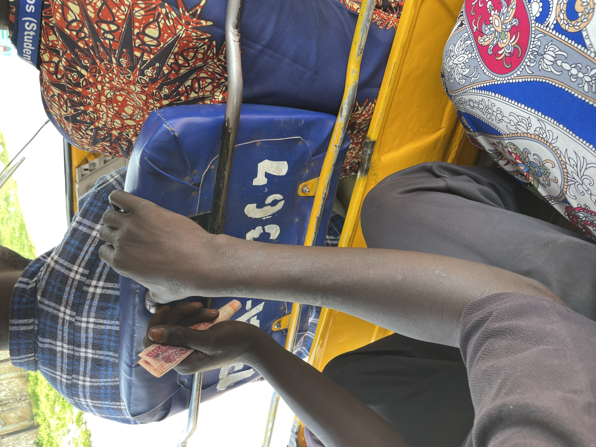 In Rafin Par, Nigeria, the image captures a close-up view of two individuals inside what appears to be a tuk-tuk, a three-wheeled motorized vehicle. One person, whose back is to the camera, is wearing a blue and white plaid shirt. Another person, facing away and to the right, is wearing a vibrant, patterned shirt with red, black, and yellow motifs. This second individual's shirt collar bears the inscription "Os (Student)".
The hands of the person in the plaid shirt are visible, holding a small wad of pink banknotes, possibly of Nigerian currency. The tuk-tuk's interior features blue padded seats and a bright yellow frame. White stenciled numbers, "367", are visible on the blue seat. The scene suggests a moment of transaction or travel within the bustling environment of Rafin Par.