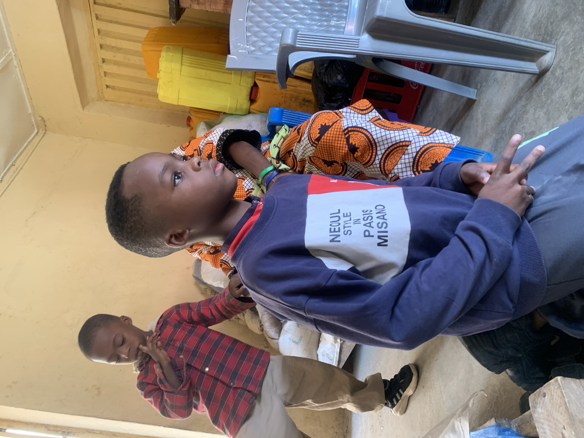 The image depicts an indoor scene, likely in Jos, Nigeria, with two young boys and a woman partially visible. The primary focus is on a boy in the foreground, wearing a navy blue sweatshirt with white lettering that reads "NEOUL STYLE IN PASIS MISANO". He is seated, looking to his right, with his hands clasped in his lap. To his left, another young boy in a red and black plaid shirt stands, looking downwards with his finger to his lips. A woman, seen from the shoulders down, is wearing a vibrant orange and black patterned fabric. The background is cluttered with various objects including large yellow and orange plastic containers, sacks, a red Coca-Cola crate, and a grey plastic chair. The walls are a light yellow, and a slatted structure is visible on the right. The lighting suggests an indoor environment with natural light entering from the left.