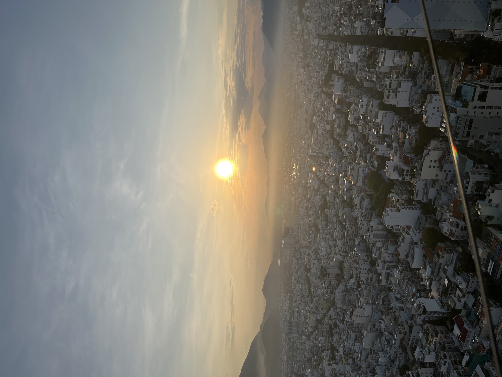 This image presents a high-angle aerial view of a densely populated urban area, likely at sunrise or sunset, in Nha Trang Ward, Vietnam. The sun, a brilliant white-yellow orb, is positioned on the horizon slightly to the left of the center, casting a bright glare and lens flare. The sky transitions from a light blue with scattered wispy clouds at the top to warm golden and orange hues closer to the horizon, indicative of a low sun angle.
A range of dark, silhouetted mountains or hills, partially obscured by atmospheric haze, spans the mid-ground, extending across the frame. A body of water, possibly a river or lake, is faintly visible winding through the landscape between the distant mountains. Below the mountains, the city unfolds with a vast expanse of tightly packed buildings, predominantly low to mid-rise structures with light-colored facades and darker roofs. Streets and thoroughfares create a grid-like pattern within the urban sprawl.
Dense urban development dominates the foreground and mid-ground, with individual buildings and blocks clearly discernible. Some greenery and trees are scattered throughout the cityscape. The overall atmosphere is hazy, particularly over the city and near the horizon, giving the scene a soft, diffuse quality. In the immediate foreground, a dark railing or structural element is visible, indicating the viewpoint is from a high-rise building or elevated platform. A building with the name "SEAESTA" is partially visible at the bottom left, and a rainbow-like light reflection appears on the railing structure. No individuals or specific activities are identifiable due to the distance and scale.
