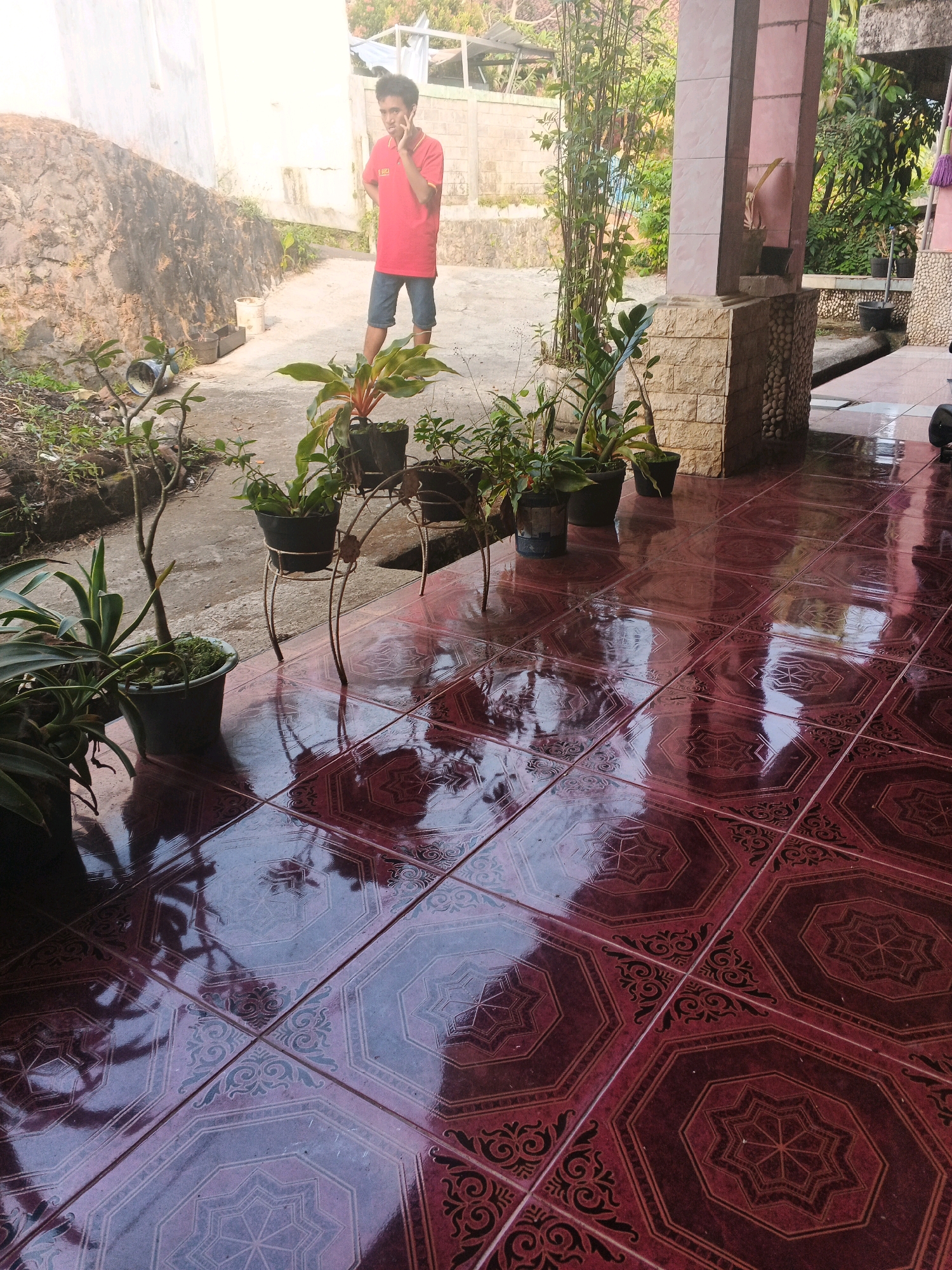The image depicts an outdoor scene in the city of Keji, Indonesia. In the background, a young man wearing a red t-shirt and blue shorts is standing and speaking on his phone. He appears to be outdoors in a courtyard or pathway. In the foreground and midground, several potted plants are visible. Some are placed on the ground, while others are on a decorative metal stand. The ground is covered with red tiles with a patterned design, which are wet and reflecting the surroundings. A building with a pink tiled column is also visible on the right side of the image, suggesting a porch or covered area. The environment appears to be a residential or semi-rural setting with some vegetation and a stone wall in the background.