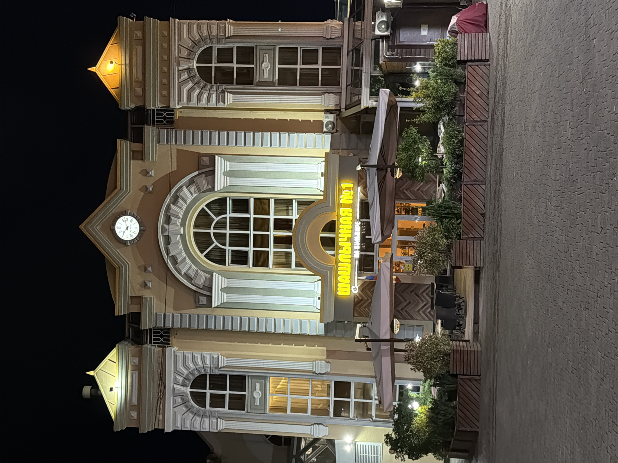 The image shows a two-story building at night, illuminated by exterior lights. The building has a grand facade with arched windows and decorative columns. Above the central entrance, a neon sign in Russian reads "ШАШЛЫЧНАЯ №1" which translates to "Shashlik No. 1". The building appears to be a restaurant with outdoor seating visible in front, featuring tables, chairs, and umbrellas. Planters with greenery line the walkway. A clock is visible on the upper part of the building. The street in front of the building is paved with cobblestones. To the right of the entrance, a red tarp or blanket is visible near a wooden door. The overall setting suggests a commercial establishment in a city. This building is located in Kislovodsk, Russia, as indicated by the context of the image.