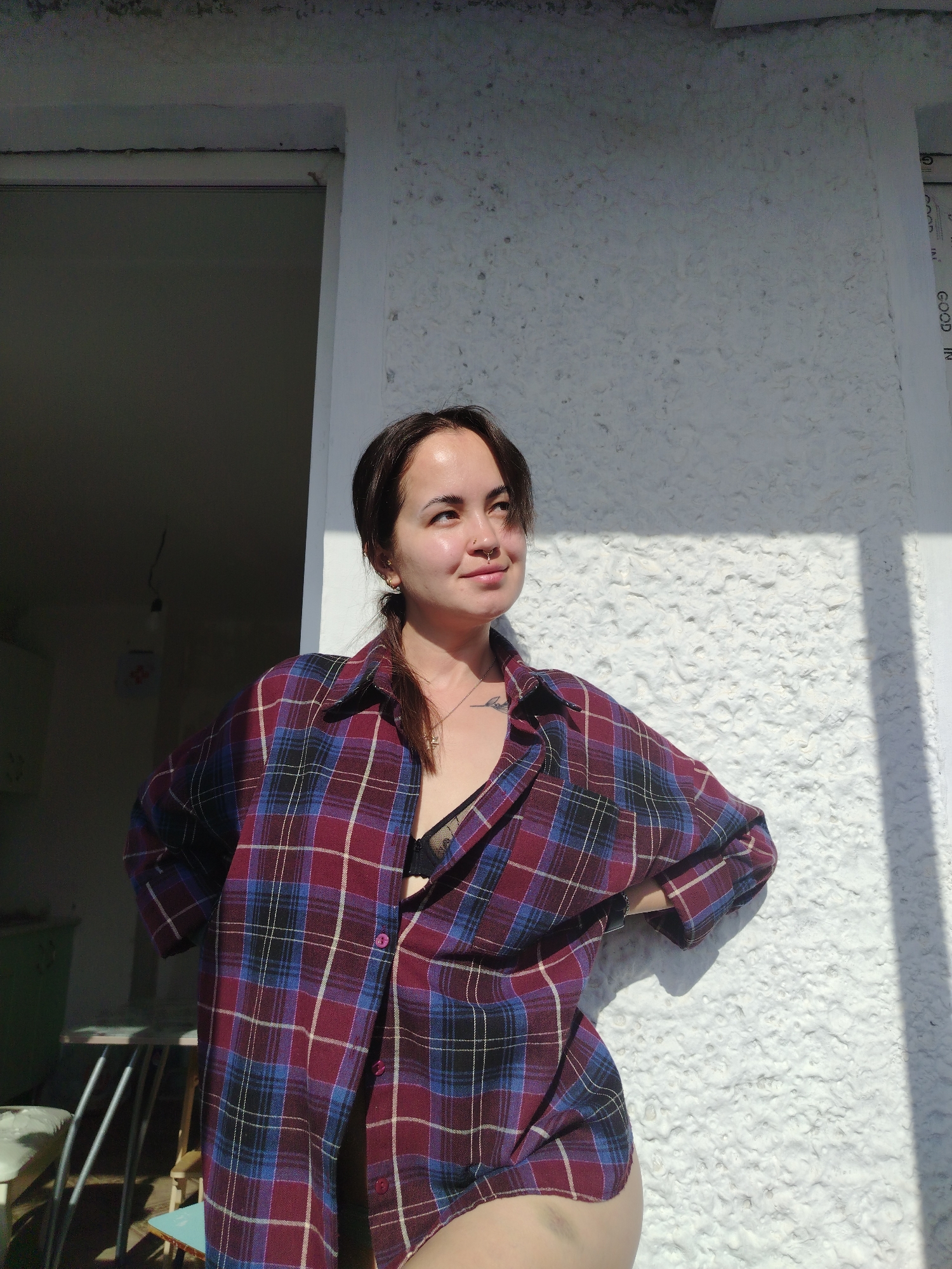 A young woman stands in a doorway or entrance, leaning against a white wall with a textured surface. She wears a large, unbuttoned, plaid shirt over a black bra. The shirt is maroon, blue, and black. The woman has dark hair, partially pulled back into a ponytail, and fair skin. A thin necklace is visible on her chest. Her face is turned to her right, and she appears to be looking upwards. The background includes a dimly lit room with furniture visible. The doorway frame is white. The image is lit with natural light, creating strong shadows. The exact location in Noviy, Russia, is not discernible from the image.