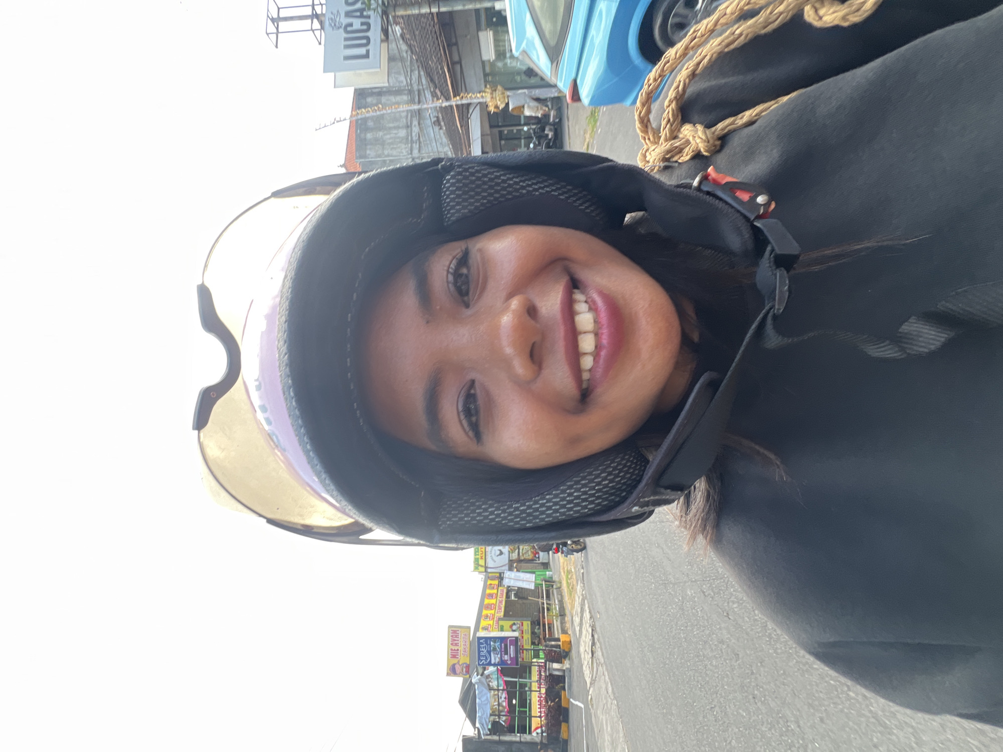 A young woman wearing a black hoodie and a helmet is taking a selfie while riding a motorcycle in Kuta, Indonesia. The woman is smiling, and her dark hair is visible beneath the helmet. In the background, there are buildings, signs with Indonesian text, and a blue car. The sky is bright and overcast. The road appears to be asphalt.