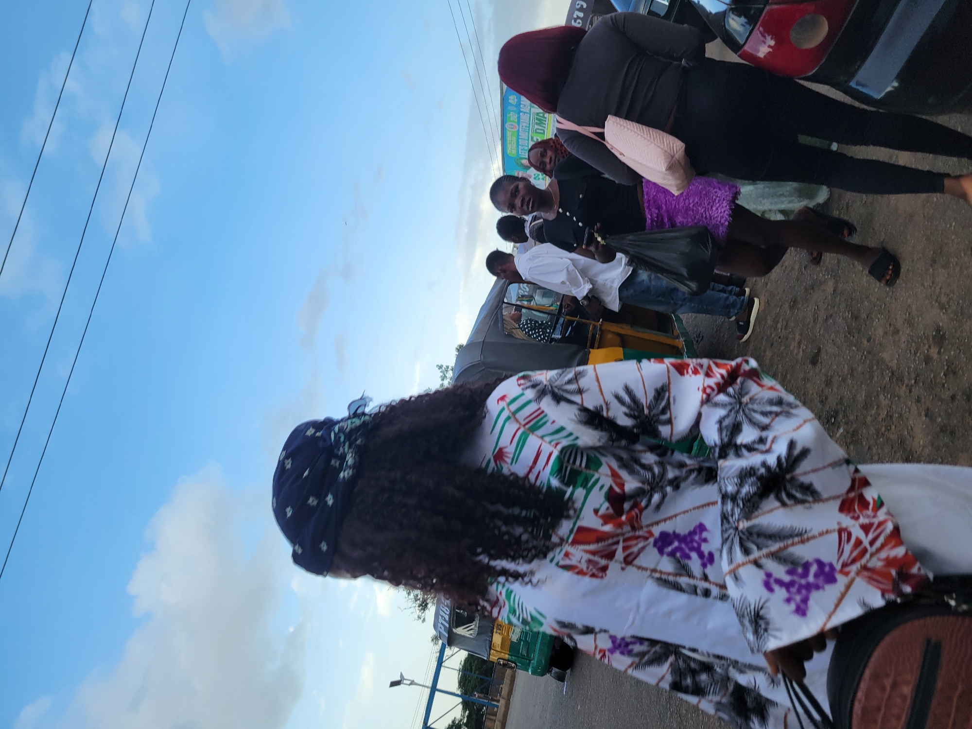 In a street scene in Jos, Nigeria, several people are gathered near a three-wheeled vehicle, likely a tuk-tuk. The sky is bright blue with scattered white clouds, suggesting it is daytime, possibly late afternoon due to the warm lighting.
A person in the foreground is wearing a white shirt with a colorful tropical pattern featuring palm trees and purple flowers, and a dark blue headscarf. Their curly hair is visible. Behind them, a group of individuals are standing around the tuk-tuk. One person, wearing a black top and dark pants, has a light pink handbag slung over their shoulder and is wearing a purple skirt. Another person is in a white shirt, and further back, a woman with reddish hair is also visible. The scene appears to be a casual street interaction or a group preparing to travel.
A sign with green and white text is visible in the background, although it is partially obscured and difficult to read. A license plate or identification number, "679", is visible on the back of a dark vehicle to the right of the frame. The ground is a mix of dirt and pavement. The overall atmosphere seems busy and typical of a street environment in Nigeria.