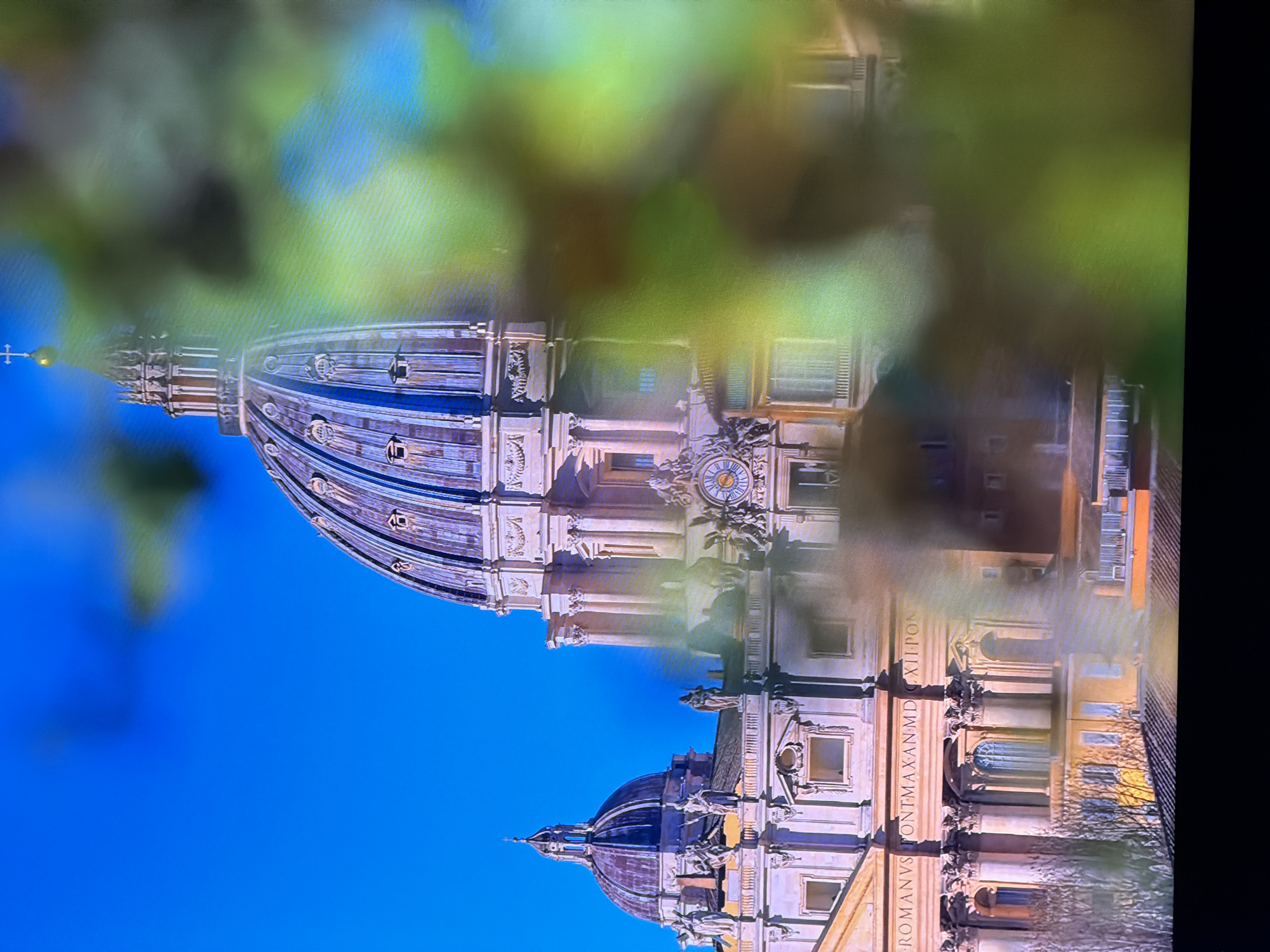 The image shows a close-up view of St. Peter's Basilica in Vatican City, Rome, Italy. The dome of the basilica is prominent in the center of the frame, with its architectural details clearly visible. Two smaller domes are also visible to the left. The building's facade, adorned with sculptures and columns, is apparent. A clock is visible on the facade of the main dome structure. The sky is a clear blue. A portion of the image is blurred by foreground foliage, which is out of focus and predominantly green, with some yellow and brown hues. There are no people or animals visible in the image. The location provided, Chirchik, Uzbekistan, is incorrect as the image depicts St. Peter's Basilica in Vatican City.