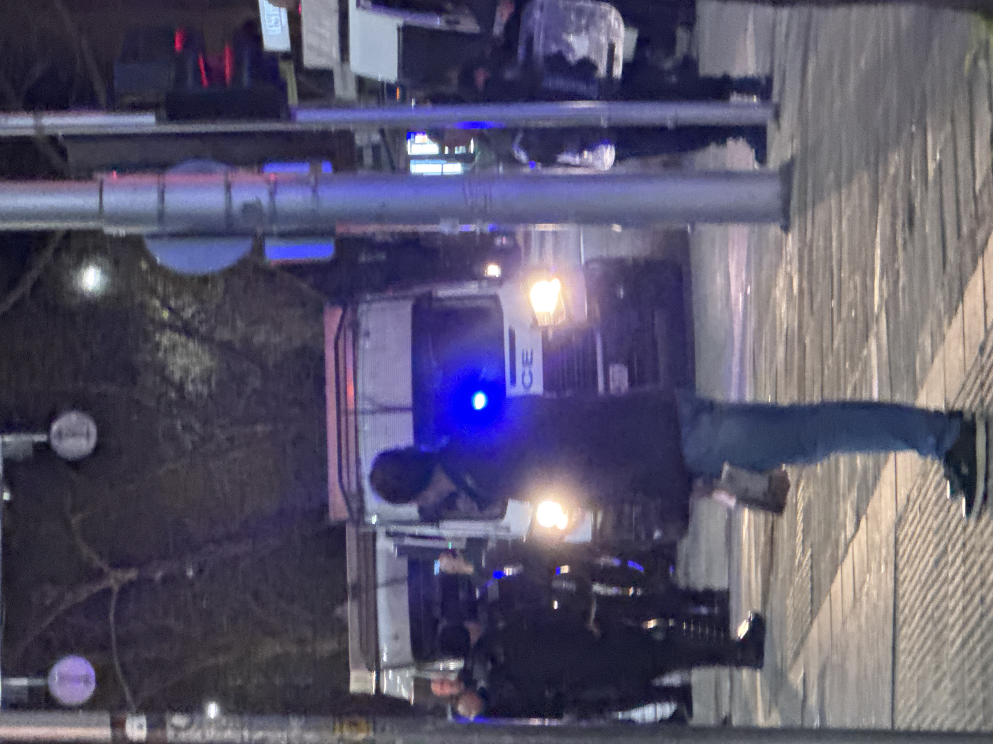 A low-angle, nighttime photo depicts a police presence at an unidentified location in Plovdiv, Bulgaria. In the midground, a police truck is parked. The word "ПОЛИЦИЯ" (POLICE) is visible on the truck's front. Several uniformed police officers stand near the truck. A civilian is standing nearby. The environment suggests an urban setting with trees and streetlights. The presence of police vehicles and officers indicates a potential public safety concern or security activity.