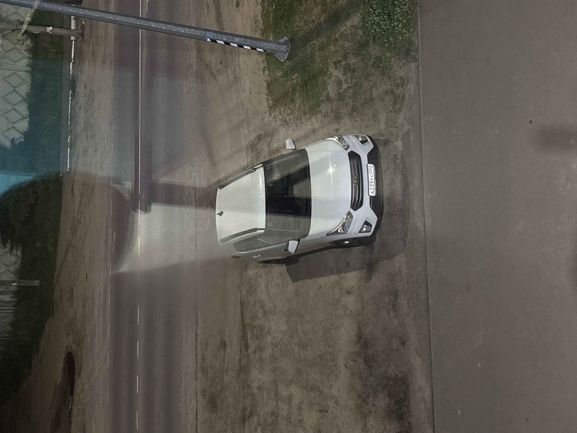 This is an overhead shot captured at night or very late dusk, showing a white Hyundai Creta car parked or stationary on a wet, dark asphalt surface. The scene is illuminated by artificial light, likely from a streetlamp, which casts a prominent bright beam across the road and the car, creating strong reflections on the damp ground.
The car is positioned towards the center-right of the frame, facing right. Its front license plate is clearly visible, displaying the Russian registration "A235YC252". Below the plate, the "HYUNDAI" logo can be faintly discerned. To the left of the car is a road with faint white lane markings visible through the light beam and reflections. Further to the left, there's a shimmering reflection of what appears to be a fence or railing.
A street lamp post, adorned with black and white stripes, stands in the upper-left quadrant of the image. Between the road and the area where the car is parked, there's a narrow strip of uneven ground with sparse grass and dirt. The overall setting is an urban or suburban street or parking area in Красные Баки, Russia, characterized by its dim lighting, wet surfaces, and the specific type of vehicle. No people or significant activity are visible, suggesting a quiet moment.