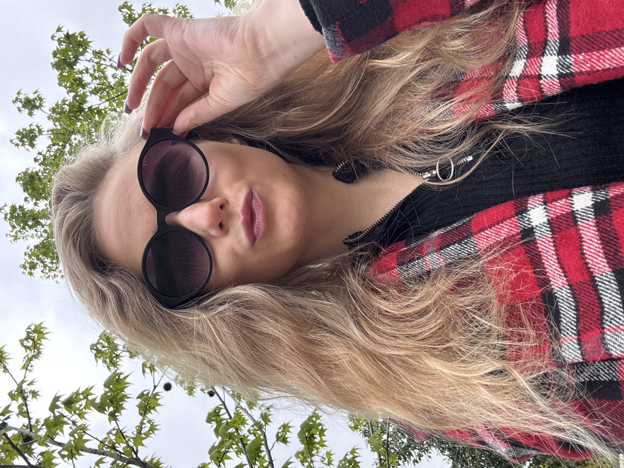 The image shows a woman with long, blonde, wavy hair wearing large, round, dark sunglasses and a red and black plaid jacket over a black, zippered top. She is holding the sunglasses on her face with her hand. The nails on her hand are painted purple. The background includes green trees and a cloudy sky, suggesting an outdoor environment in the city of Unknown, Unknown. The camera angle is low, looking up at the subject.
