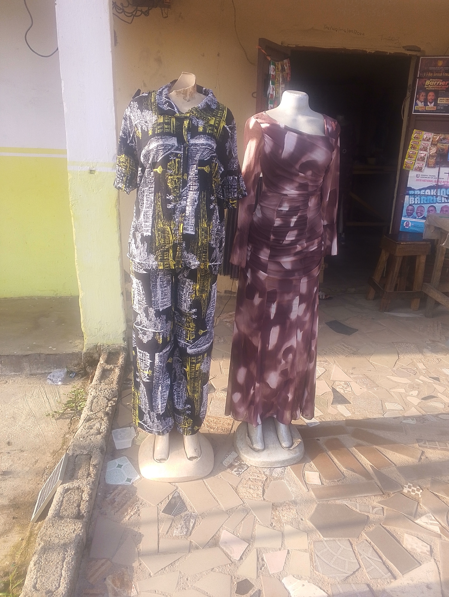 This is an eye-level, medium shot of two mannequins standing outside a shop in Jalingo, Nigeria. The mannequin on the left is wearing a black and yellow patterned shirt and pants, while the mannequin on the right is wearing a long, patterned brown dress. The shop entrance is visible behind the mannequins, along with posters and shelves stocked with goods. The ground in front of the shop is paved with irregular tiles, and there is a wall to the left of the mannequins. The scene appears to be outdoors, during daylight hours.