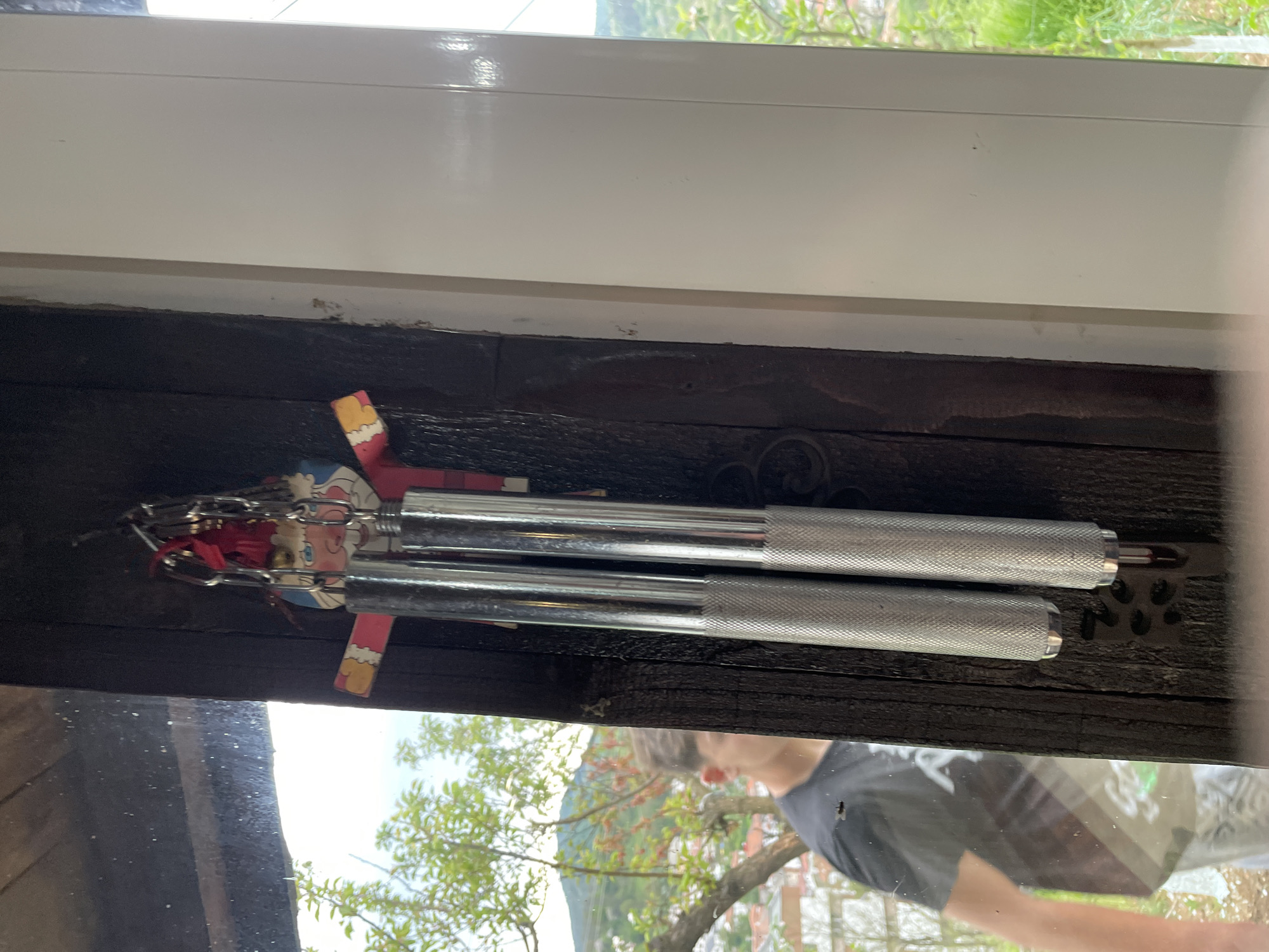 Two silver-colored objects with knurled grips are lying horizontally on a dark wooden surface. Attached to one end of the upper object is a small figure with a chain and red material. The objects are located below a window with a white frame. A reflection in the window shows a person with short brown hair wearing a gray shirt, along with a tree and foliage. The location of the image is unknown and cannot be determined as Zlatograd, Bulgaria.