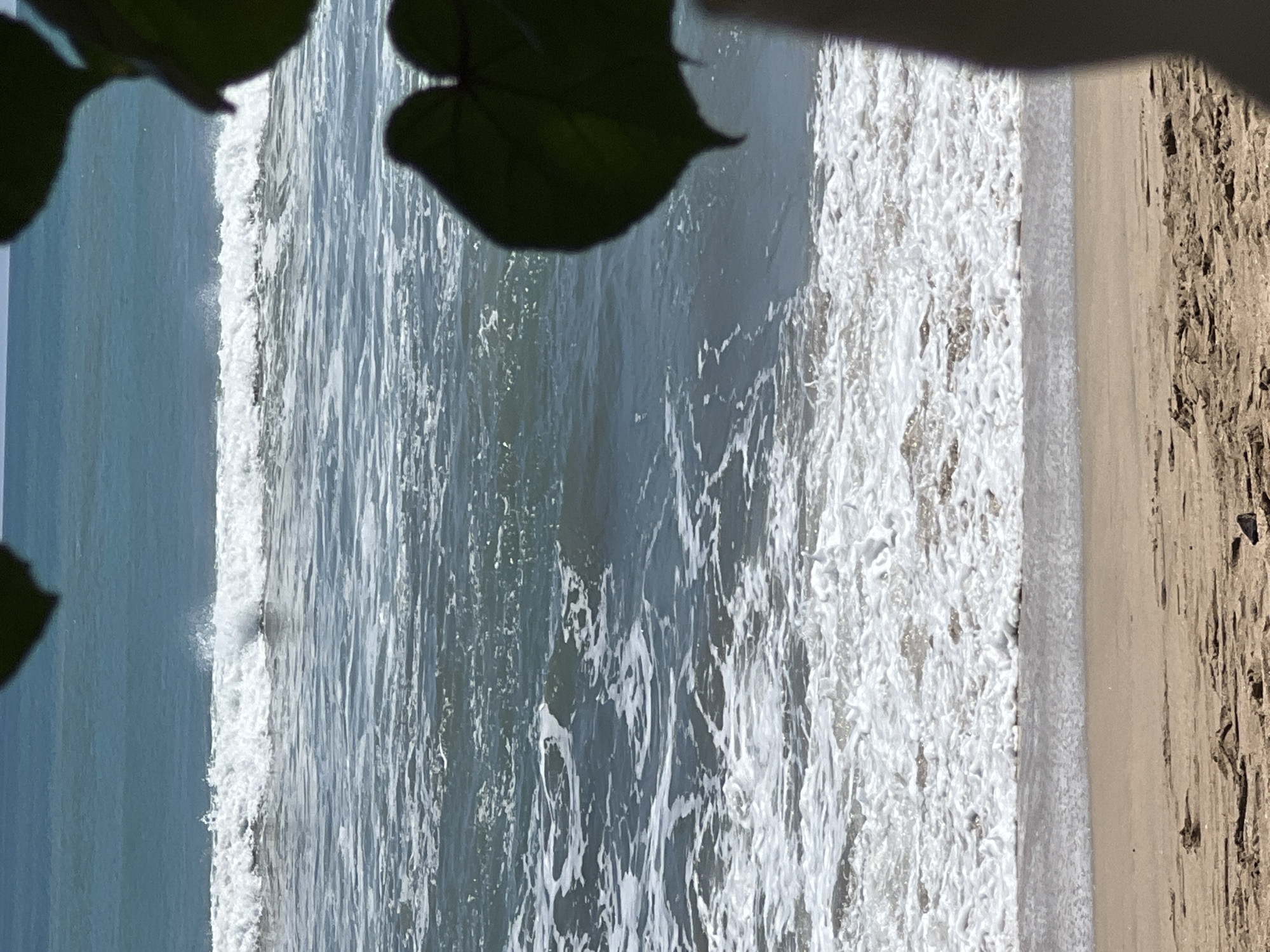 A tranquil ocean scene unfolds in Kuta, Indonesia, with gentle waves lapping at a sandy shore. The water, a blend of blues and greens, is dotted with white foam as waves break near the beach. The foreground reveals the texture of wet sand, with a few scattered debris. In the upper right corner, a large, dark green leaf frames the view, hinting at the lush vegetation often found near Kuta's coast. The sky above is a soft, hazy blue, suggesting a warm, sunny day. The overall impression is one of peaceful serenity, a typical coastal vista of Kuta.