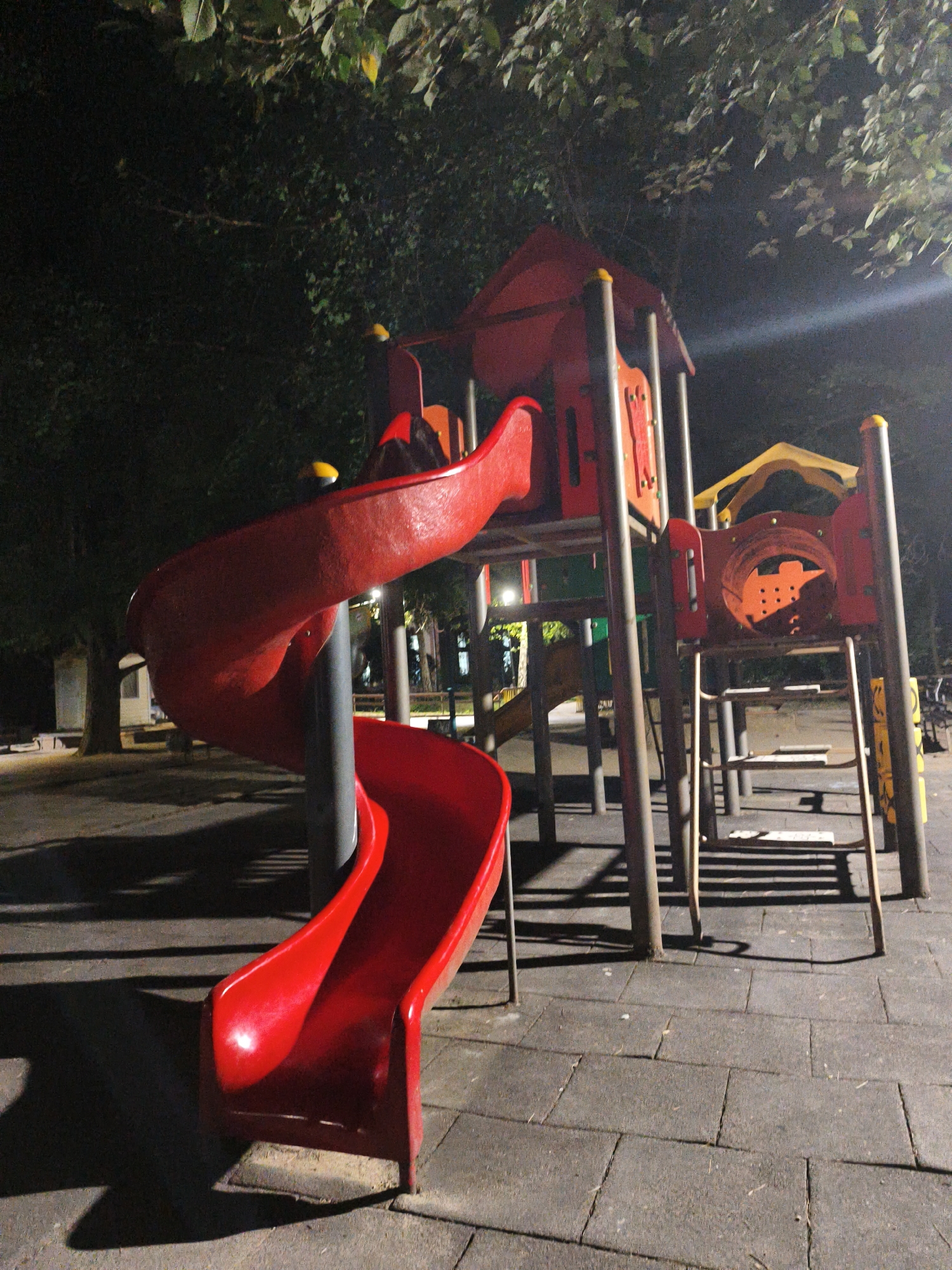 This image depicts a brightly lit red spiral slide at a children's playground, viewed from a low angle at night. The slide is the prominent feature, with its smooth, curved surface catching the artificial light. The playground structure is made of metal and plastic, with other red and yellow elements visible. The ground is paved with dark square tiles, and long shadows are cast across the surface, indicating a strong light source. In the background, trees with lush green leaves are visible, silhouetted against the dark night sky. The lighting suggests the playground is located in a park or outdoor recreational area. The specific location in Lovech, Bulgaria is not explicitly indicated by any visual cues within the image.