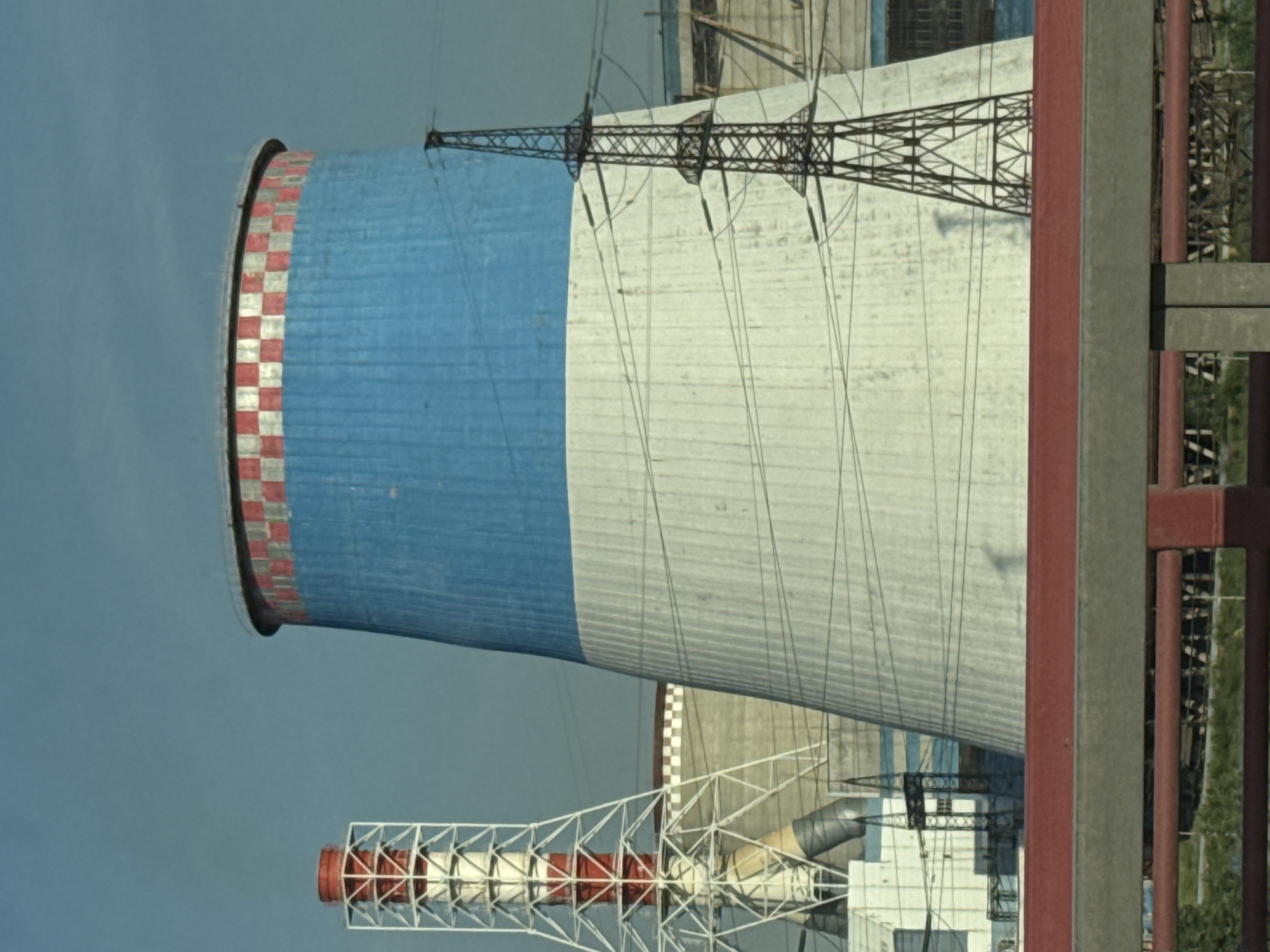 The image shows a cooling tower of a thermal power station, a tall industrial chimney, and transmission towers against a hazy blue sky. The cooling tower is massive and conical, painted with a blue band near the top, with a red and white checkered pattern around its rim. The chimney on the left is a lattice structure with red and white elements. Power lines are strung between transmission towers. In the foreground, a red railing and a concrete barrier are visible, suggesting the image was taken from a bridge or elevated walkway. In the background, other industrial structures and buildings are partially obscured. This scene is likely from the southern part of Saint Petersburg, Russia, where several large power plants are located. There are no people or animals visible in the frame.