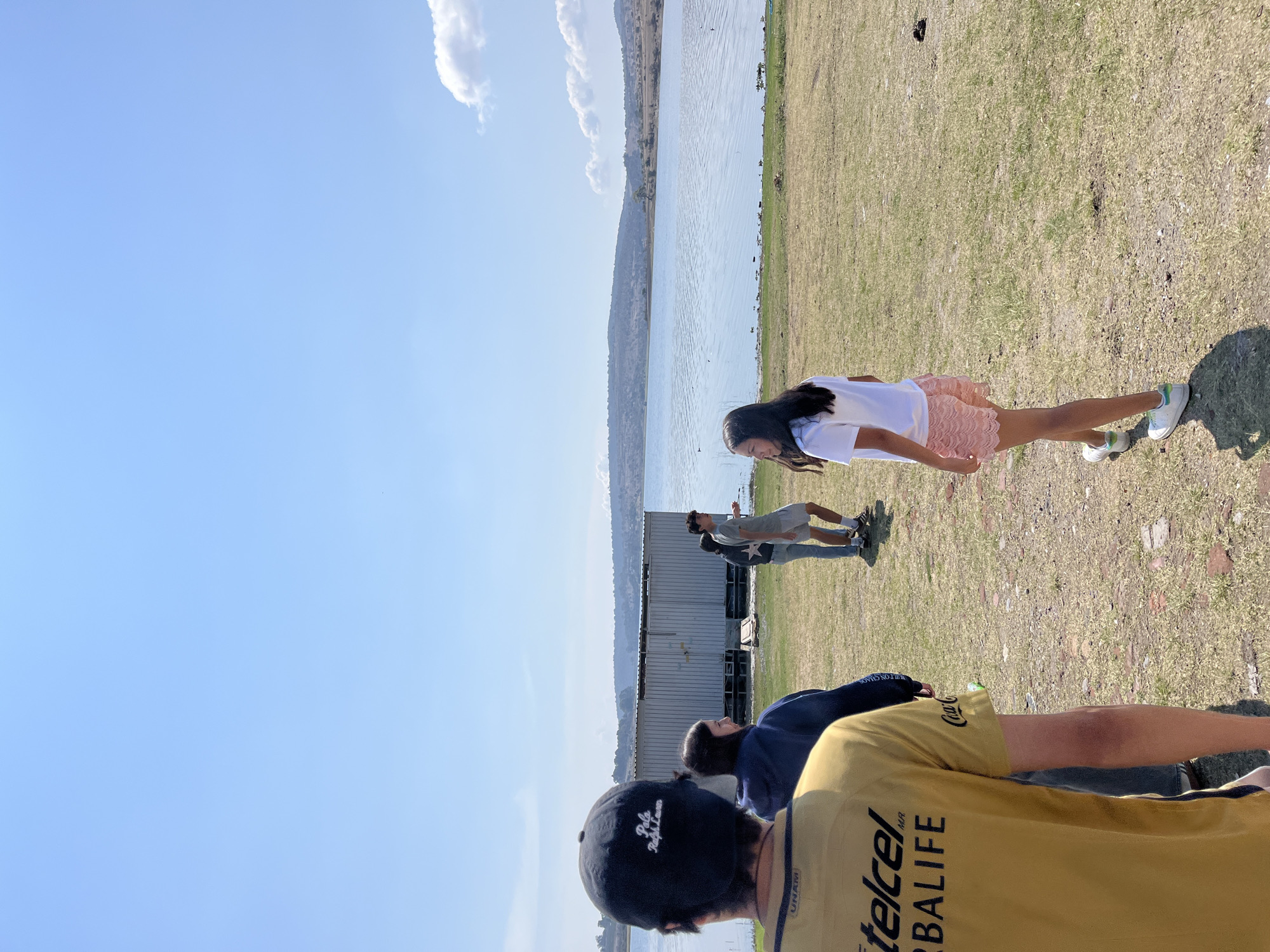 The image depicts an outdoor scene in El Oasis Valsequillo, Mexico. A body of water borders a grassy area where several people are present. A young woman wearing a white shirt and pink skirt stands in the foreground. Behind her, a man in a grey shirt and shorts stands near a silver building. In the foreground, a person wearing a black baseball cap and a yellow shirt are seen from the back. Another person wearing a blue jacket is also partially visible. The sky is blue with a few clouds.