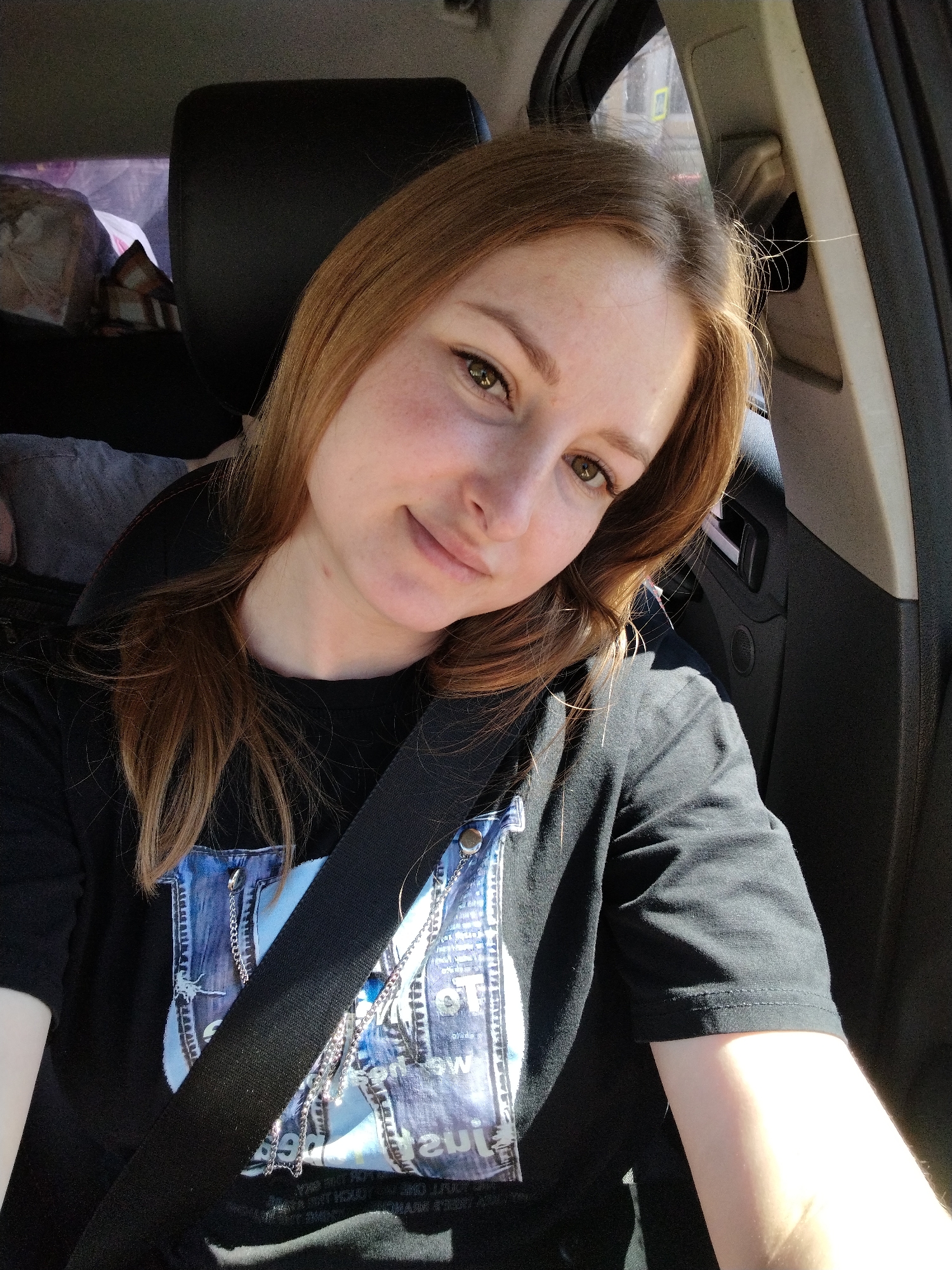 A light-skinned woman with auburn hair is sitting in the driver's seat of a car, wearing a black t-shirt with a blue and white graphic design and a seatbelt across her chest. She is looking directly at the camera with a slight smile. The car's interior is visible, including the headrest of the seat behind her and the car door. The location is possibly in Kazan, Russia, but there is no definitive evidence of this in the image. The environment suggests it is daytime.