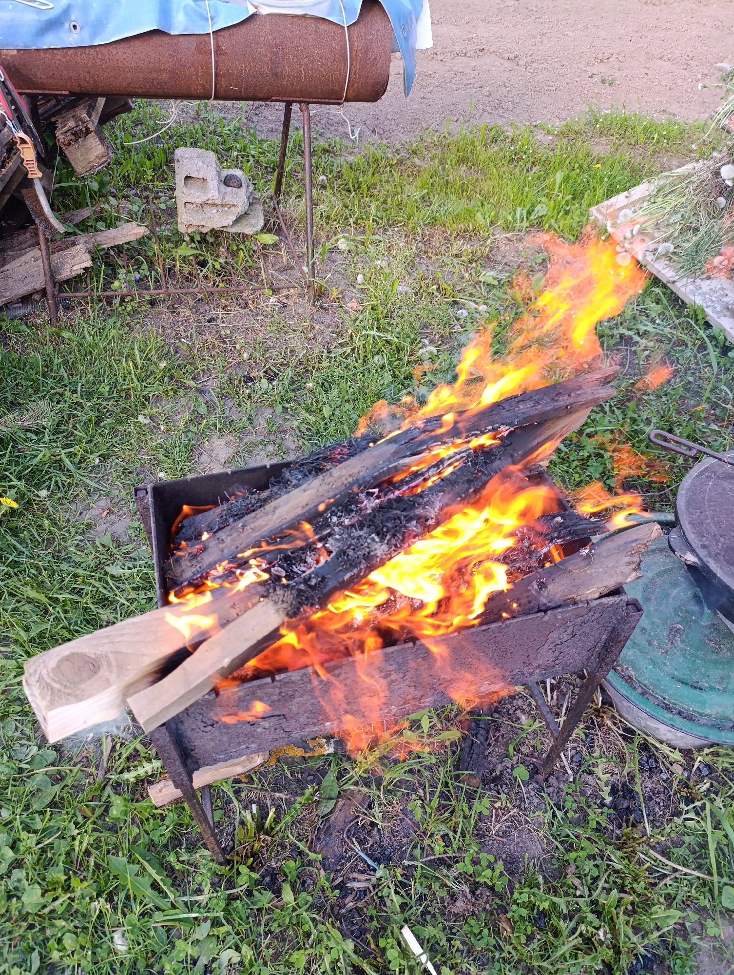 The image shows a burning pile of wood in a metal fire pit with stand legs on a grassy ground. The wood is a mix of charred and fresh pieces, with flames rising high. A metallic barrel and concrete block are visible in the background. It is an outdoor scene during daylight.