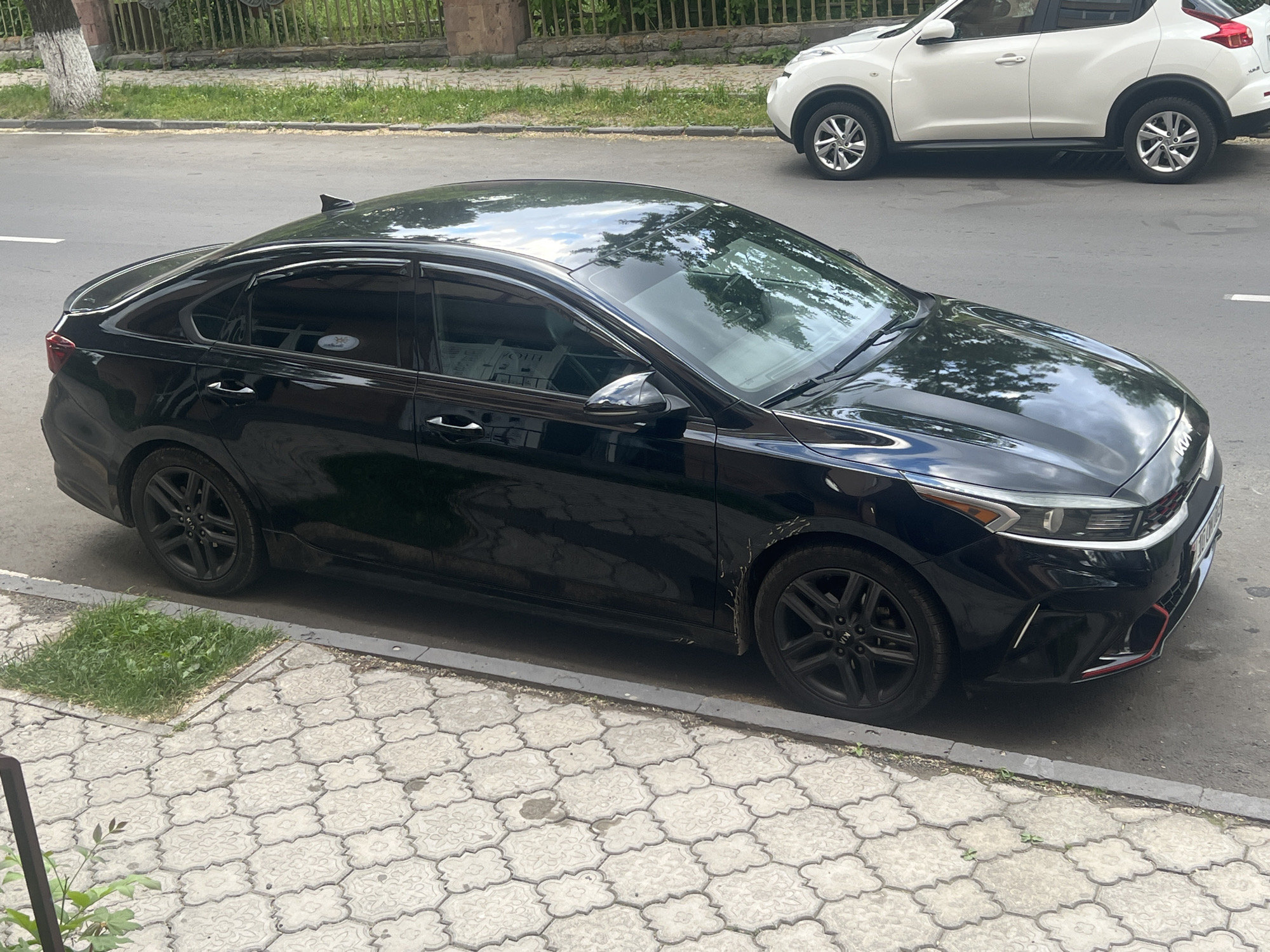 The image shows a black sedan parked on the side of a street. The car is the primary subject. The street is paved, and there is a sidewalk with decorative grey paving stones next to it. Some patches of grass are growing through the paving stones and at the edge of the road. In the background, there is a white SUV also parked on the street, and behind that, a low stone wall and some greenery. The scene appears to be set outdoors during the daytime. The lighting suggests it might be a sunny day, as evidenced by reflections on the cars. The overall atmosphere is ordinary, depicting a typical urban or suburban street scene. There are no people visible in the image. The car's make is partially obscured, but the logo on the wheel appears to be "KIA". There is some visible dirt or scuff marks on the lower body of the black car.