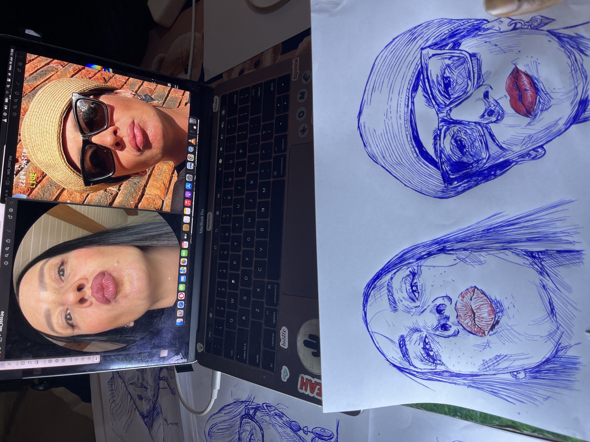 The image shows a close-up view of a laptop screen displaying two video call participants. On the left, a woman with long dark hair puckers her lips. On the right, a woman wearing a straw hat, sunglasses, and a beret also puckers her lips. The laptop is open and placed on a surface, likely a desk, where two blue ink drawings of women's faces are laid out in front of it. One drawing is of a woman with a similar pose to the one on the left of the screen, and the other drawing depicts a woman with glasses and a beret, mirroring the woman on the right of the screen. The drawings appear to be in progress. There is no identifiable indication of the location being Jos, Nigeria.
