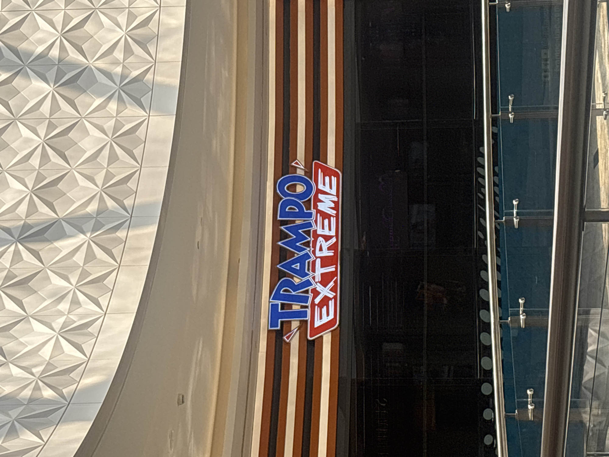 The image captures a sign for "Trampo Extreme" prominently displayed on the facade of a building. The sign features blue lettering for "TRAMPO" and red and white lettering for "EXTREME," set against a background of horizontal stripes in varying shades of brown, cream, and white. Below the sign, a dark, reflective glass storefront is visible. The architectural style of the building includes a curved ceiling with a patterned, textured white surface. In the foreground, a modern railing system with glass panels and metal fixtures is present, suggesting this is an indoor location within a mall or entertainment venue. The lighting suggests it is daytime, with sunlight casting shadows on the patterned ceiling. This is likely in Dubai, United Arab Emirates, given the branding and typical mall architecture found in the region. There are no people clearly visible within the frame.