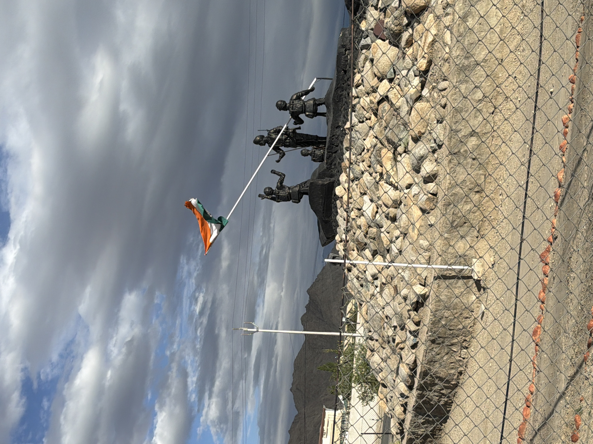 The image depicts a monument featuring several bronze statues of soldiers in an outdoor setting with mountains in the background. An Indian flag is unfurled on a pole attached to the monument. The monument is situated on a raised platform covered with rocks and stones, and a chain-link fence is in the foreground. A utility pole with wires is visible to the left of the monument. The sky is cloudy. The scene is located in Spituk, India.