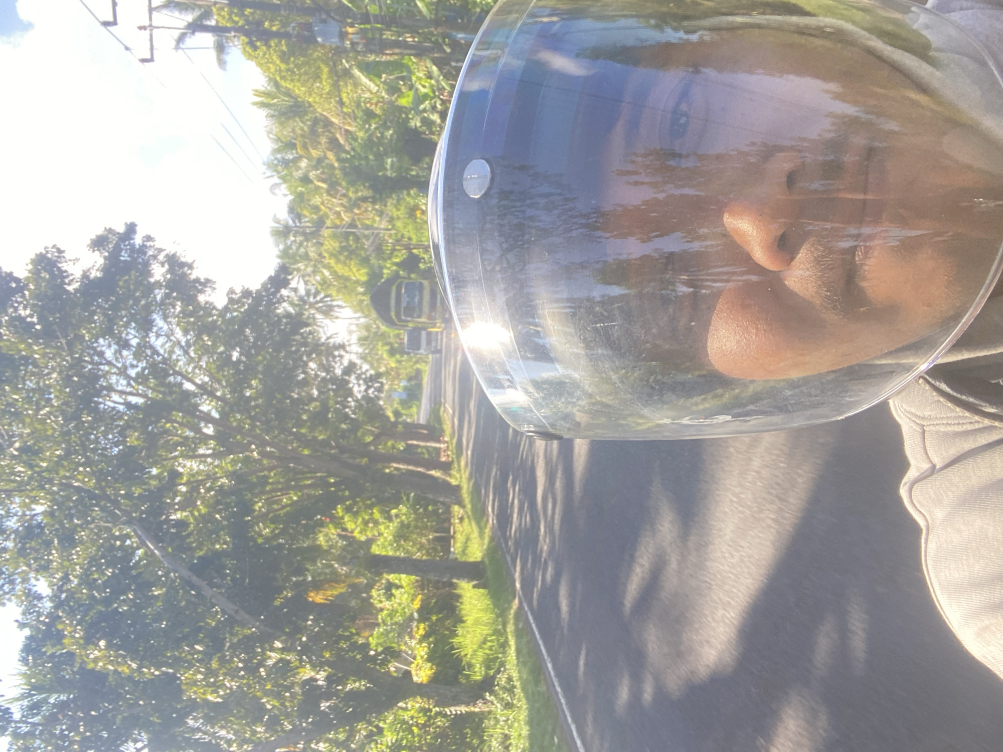 The image depicts a first-person perspective from a motorcycle traveling on a paved road in Selemadeg Barat, Indonesia. The road is lined with lush green grass and tall trees with dense foliage, casting shadows on the asphalt. Sunlight filters through the trees, creating a bright and slightly hazy atmosphere. In the distance, two trucks and a white van are visible on the road. The foreground is dominated by the rider's helmet, with a clear visor reflecting the surrounding environment and the rider's face. The rider appears to be a male with a mustache, looking directly ahead. The overall scene suggests a bright, sunny day in a tropical or subtropical rural area.