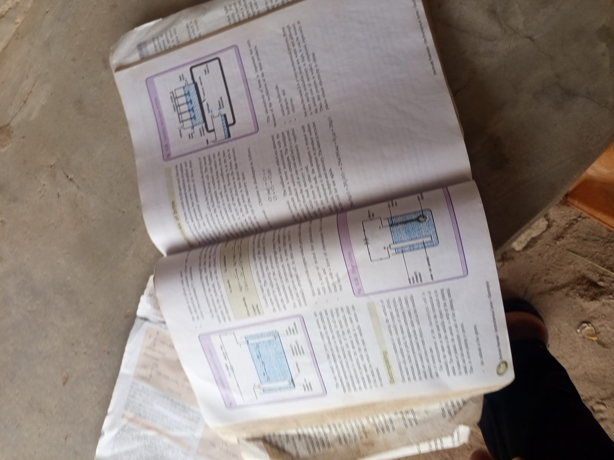 An open textbook lies on a sandy, textured surface that appears to be the ground in Jalingo, Nigeria. The book's pages, filled with text and diagrams, are slightly crinkled. One page shows a diagram of an electrolytic cell with labels like "anode," "cathode," and "electrolyte." Another section of the text discusses the electrolysis of brine. The lighting suggests it is daytime.
